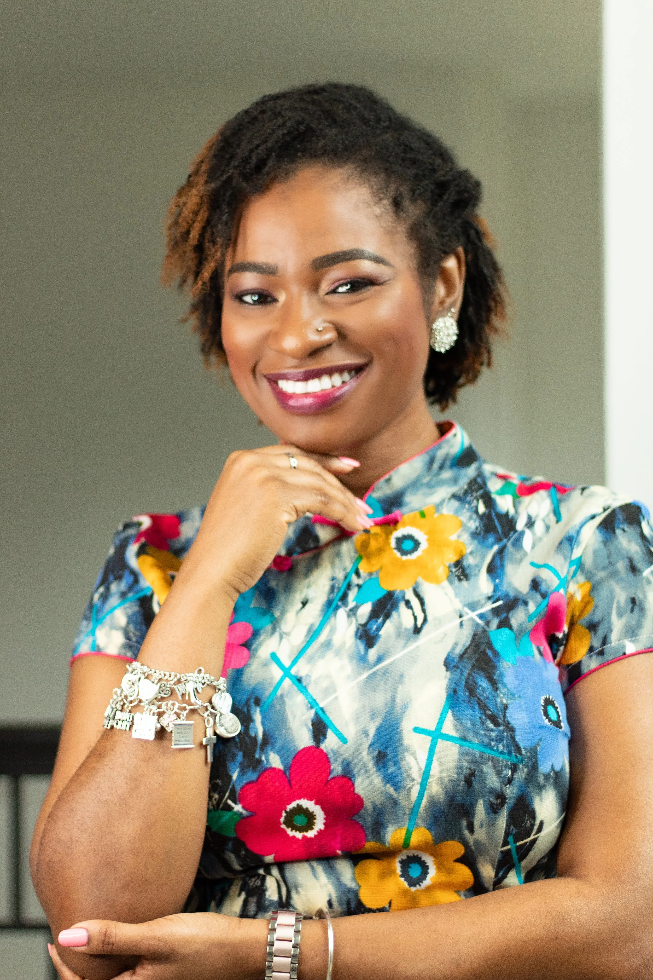 Close-up of a smiling woman with dark skin, short curly hair, wearing a colorful floral dress, earrings, and bracelets, with her right hand resting near her chin and left arm crossed over her waist.