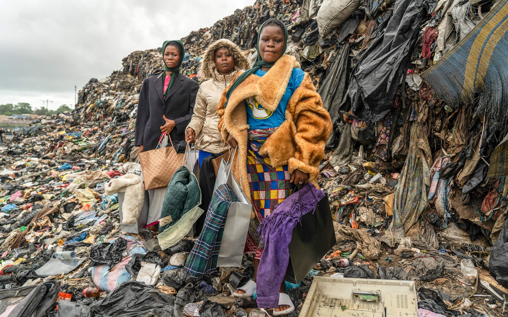 Trois jeunes filles portent des sacs de vêtements ou d'objets, debout dans un dépotoir à ciel ouvert rempli de déchets, avec un monticule de déchets derrière elles.