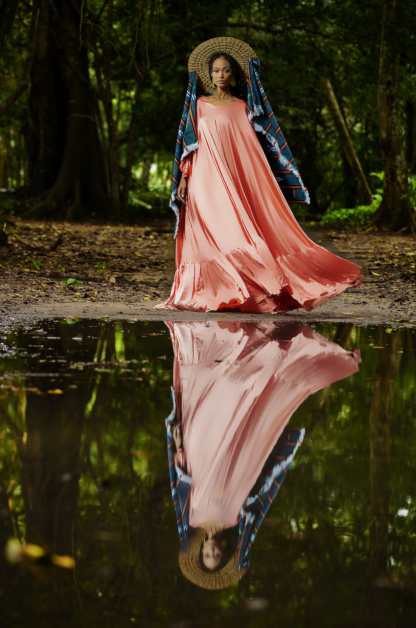 Femme en robe rose fluide, portant un grand chapeau en paille, debout dans une forêt avec son reflet dans l'eau