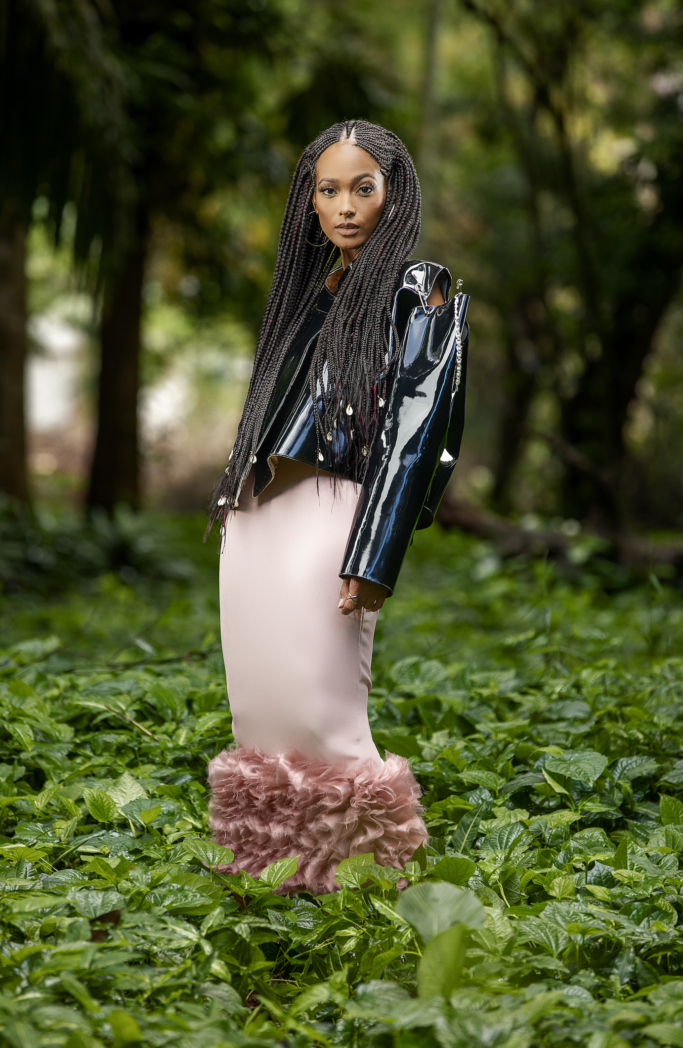 Une femme porte une veste en PVC noir brillant, une jupe longue rose pâle avec des franges en tissu au bas, et se tient dans une forêt verdoyante.