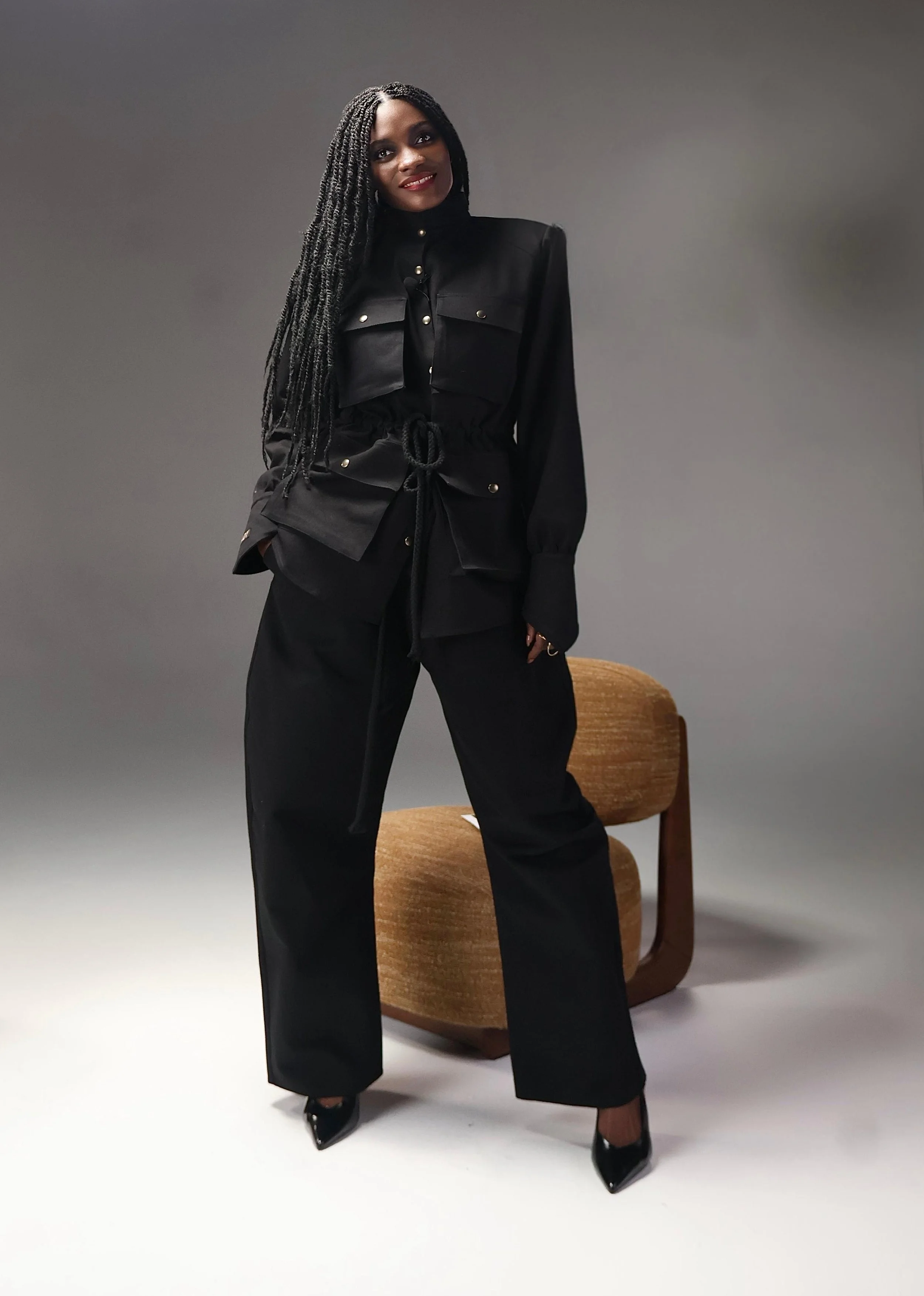 Une femme porte une tenue noire élégante avec des pantalons amples, un haut à manches longues et une veste à plusieurs poches. Elle se tient debout devant un fond gris et une chaise en bois. Elle a des cheveux longs en torsades et un maquillage rouge.