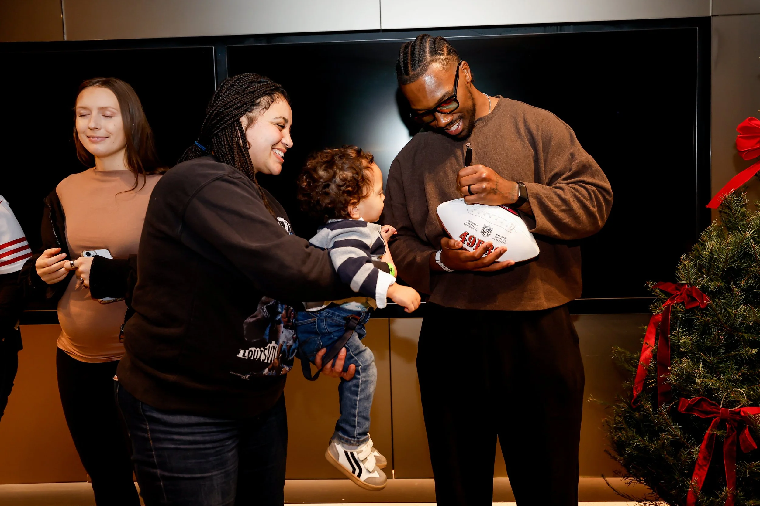 A group of people at a festive event, with a woman holding a young child, a man signing a football for the child, and a Christmas tree with red ribbons nearby.