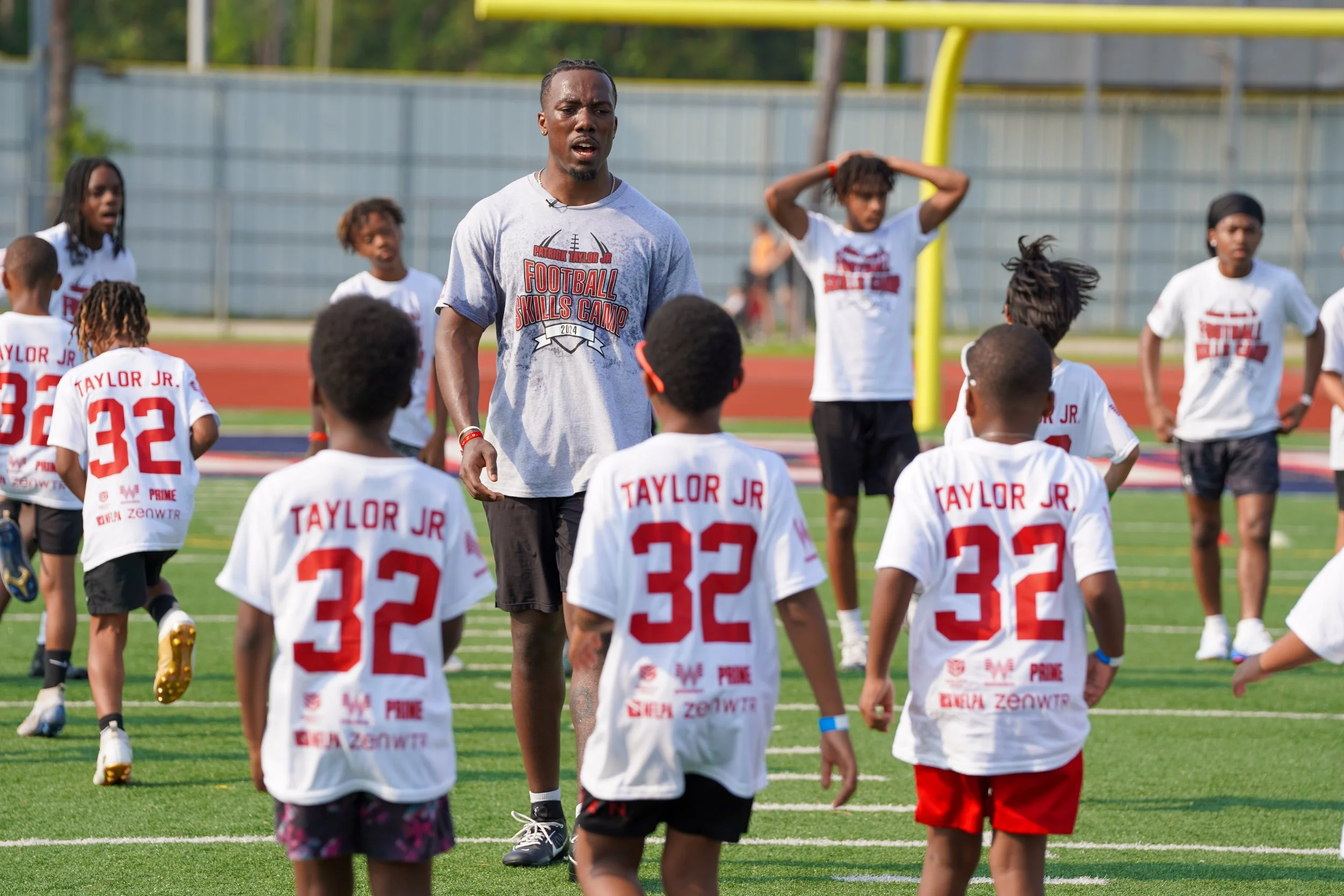 Football coach instructs young players during practice on a football field, with children wearing white jerseys labeled 'TAYLOR JR' and players listening attentively.