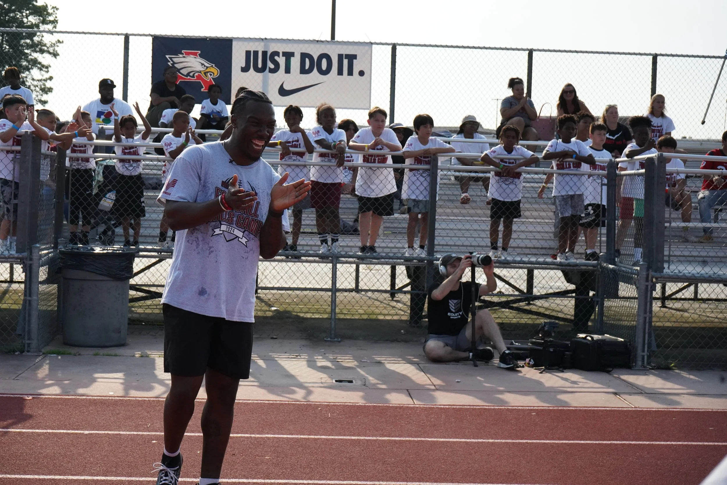 A man in athletic clothing claps and smiles on a running track, with children watching from bleachers behind a chain-link fence, and a photographer sitting on the ground nearby. There is a banner with a bird logo and the slogan 'Just Do It.' in the b