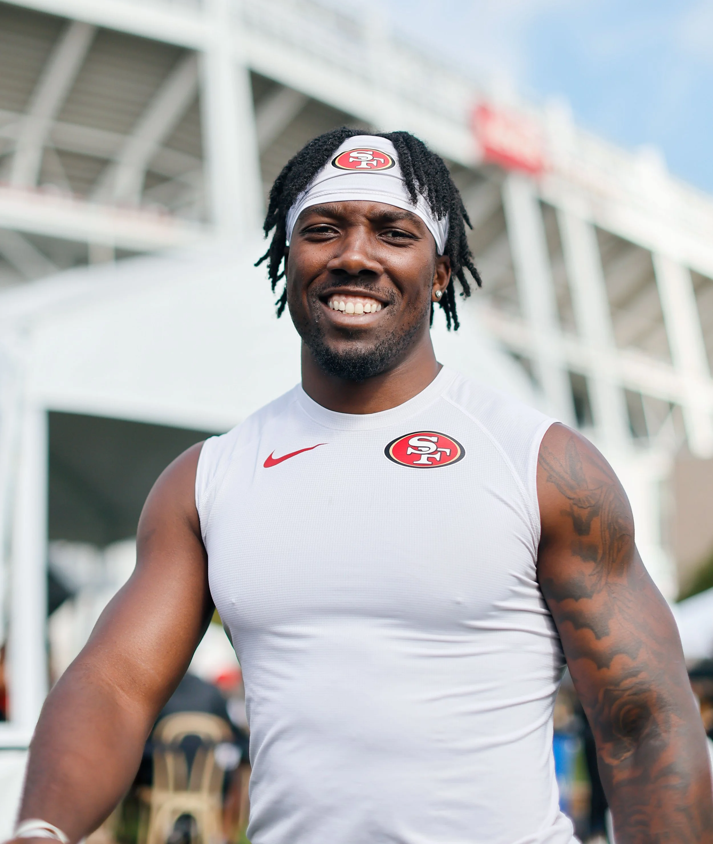 Patrick Taylor Jr. wearing a white tank top and headband with San Francisco 49ers logo, outdoors with stadium structure in the background.