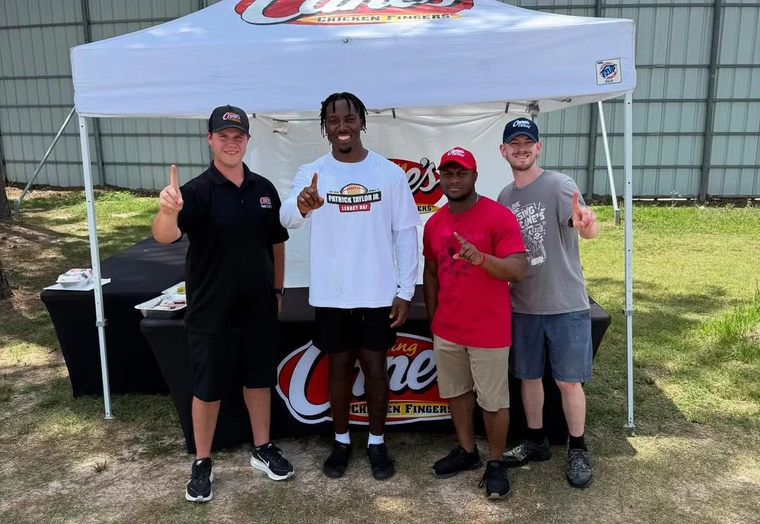 Four men standing in front of a Canes Chicken Fingers tent, smiling and posing for the camera, with three raising one finger.
