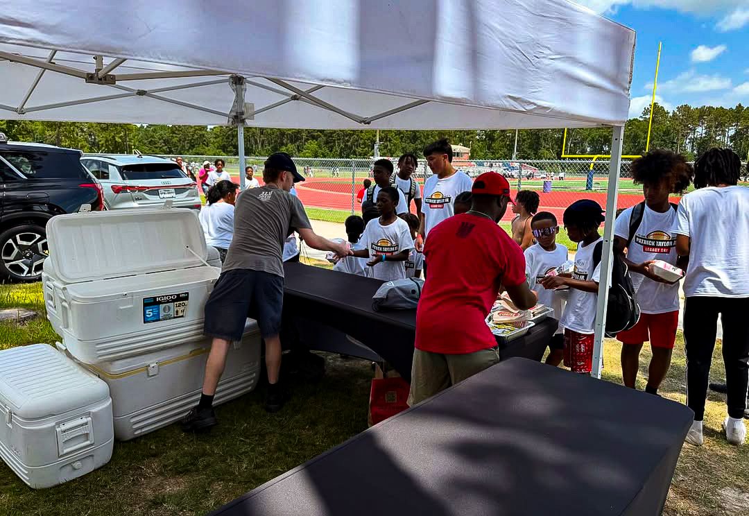 People gathered under a tent at an outdoor event, with children and adults standing in line, possibly receiving food or supplies. There are parked cars, a track, and trees in the background.