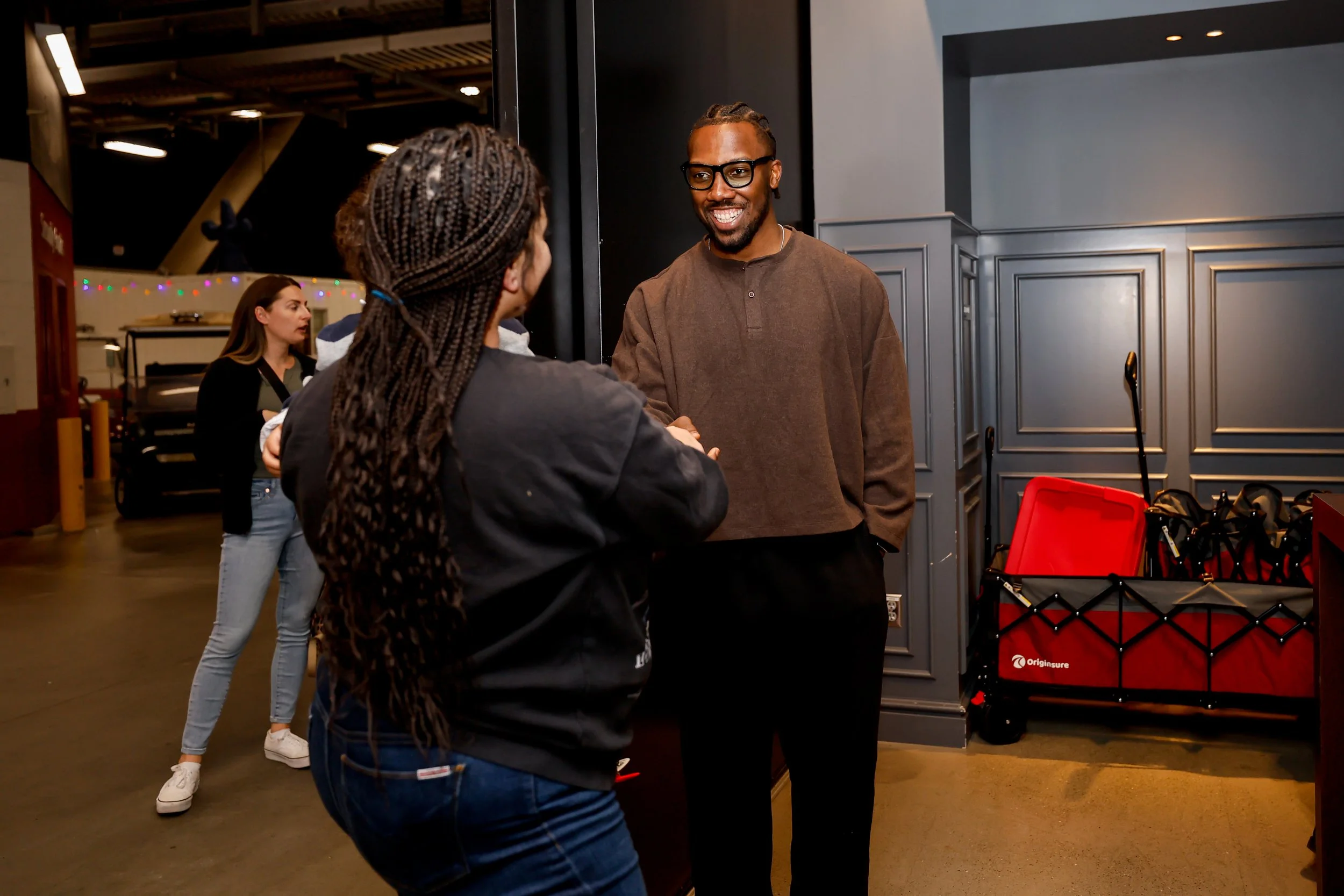 A man wearing glasses and a brown shirt shaking hands with a woman with braided hair in a black jacket inside a building.