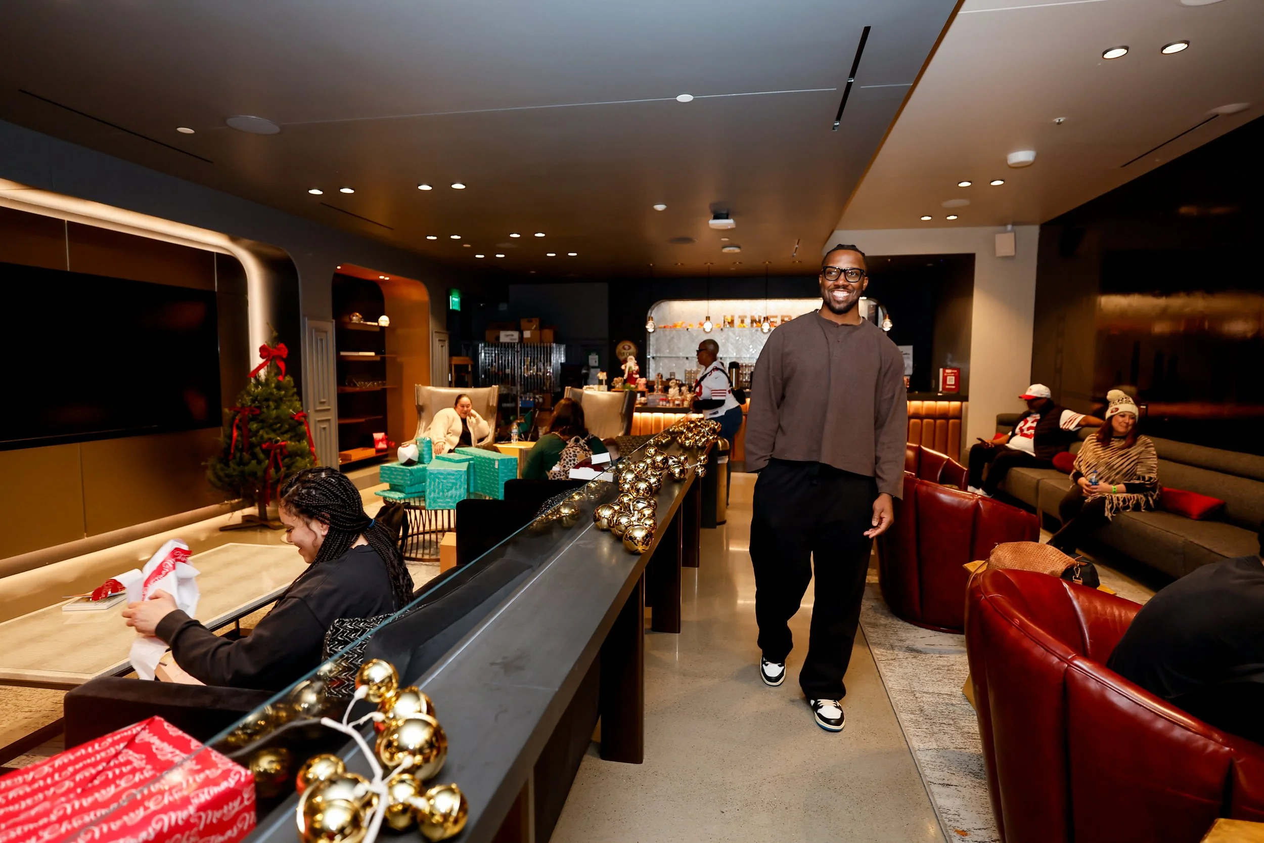 People in a cozy, decorated restaurant during Christmas. A man walking in the foreground, smiling. Christmas tree and presents on a table, holiday decorations including gold ornaments. Guests seated, some exchanging gifts, others chatting.