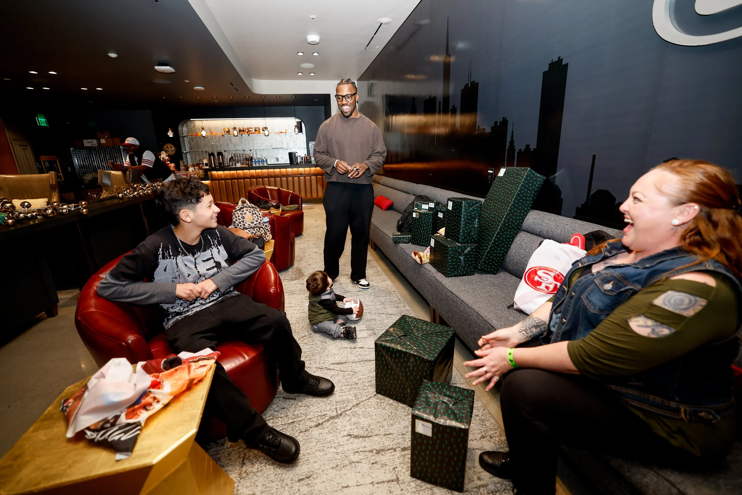 Group of people enjoying a gift exchange in a modern lounge, with wrapped presents and a bar in the background.