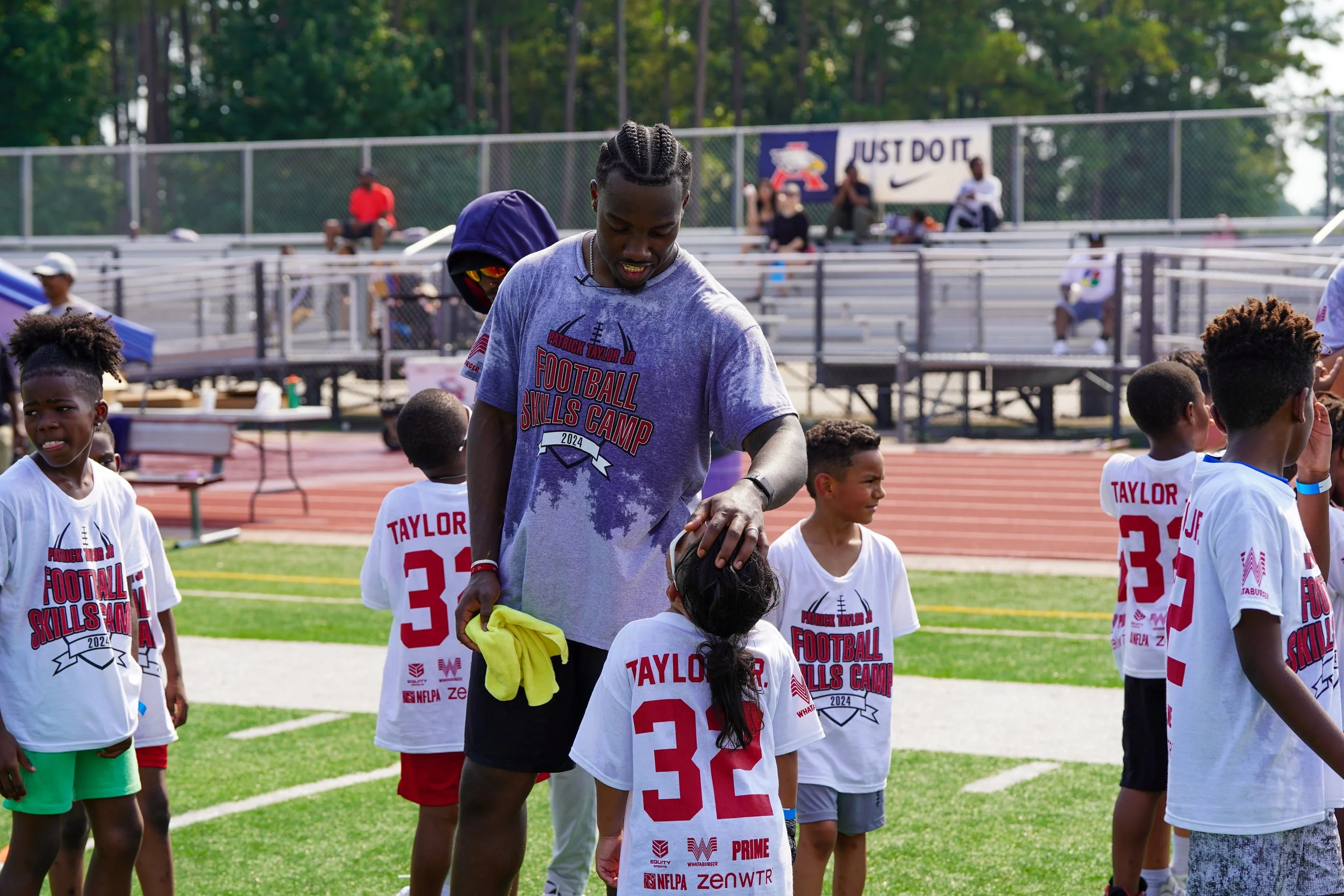 A football coach is giving instructions or encouragement to young players during a football skills camp on a field, with a few people sitting on bleachers and banners in the background.
