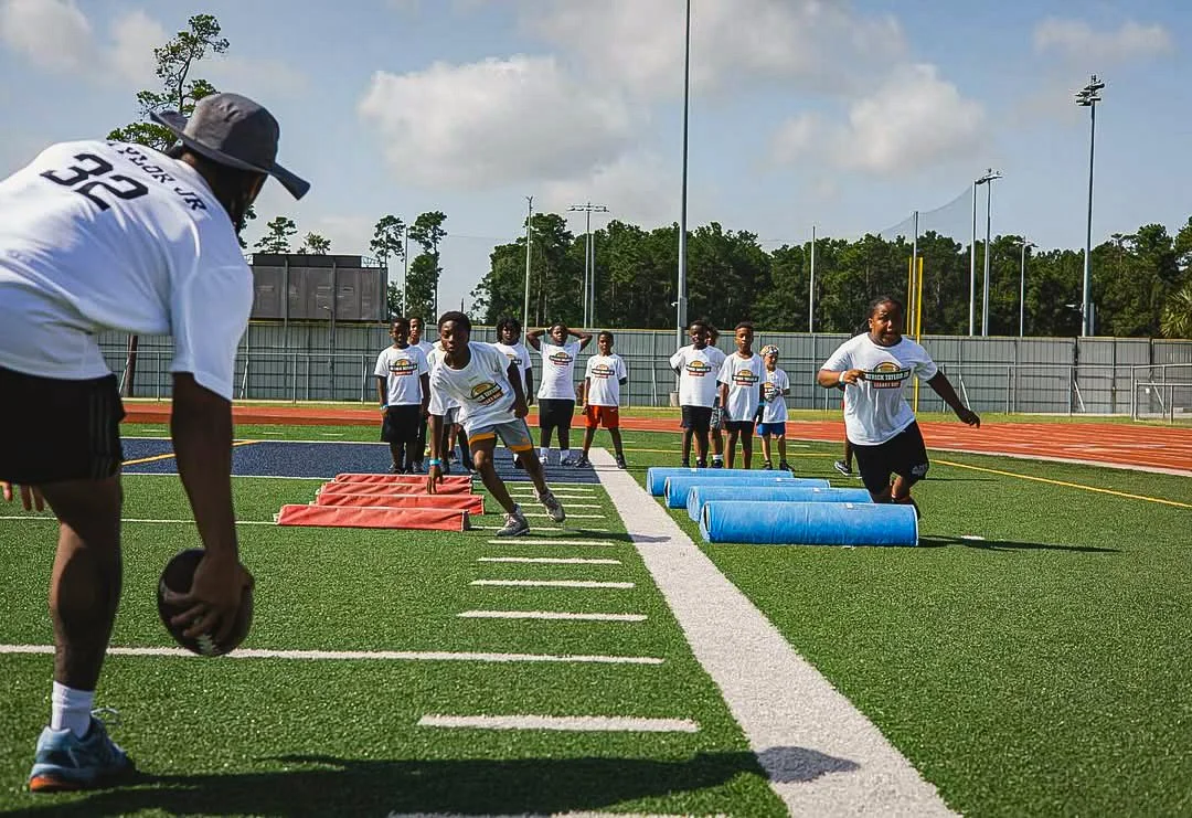 Young athletes participate in an outdoor football training session on a field with cones and agility drills, supervised by a coach with a football.