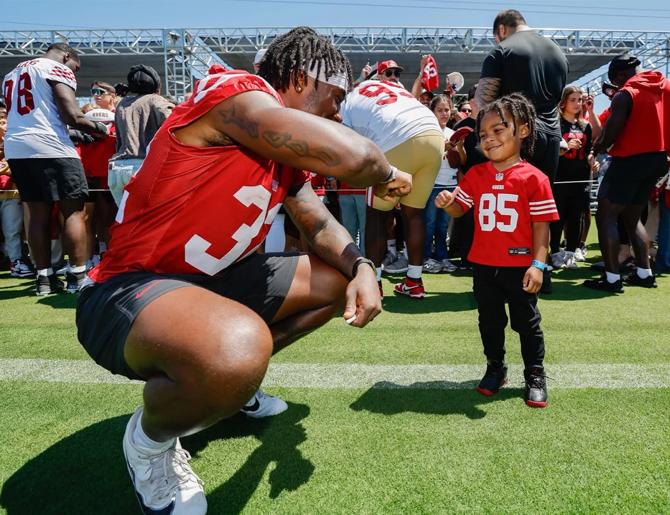 Patrick Taylor Jr. squats and bumps his fist with a young kid in a red jersey with the number 85, who is smiling. They are on a football field with a crowd and stadium seating in the background.