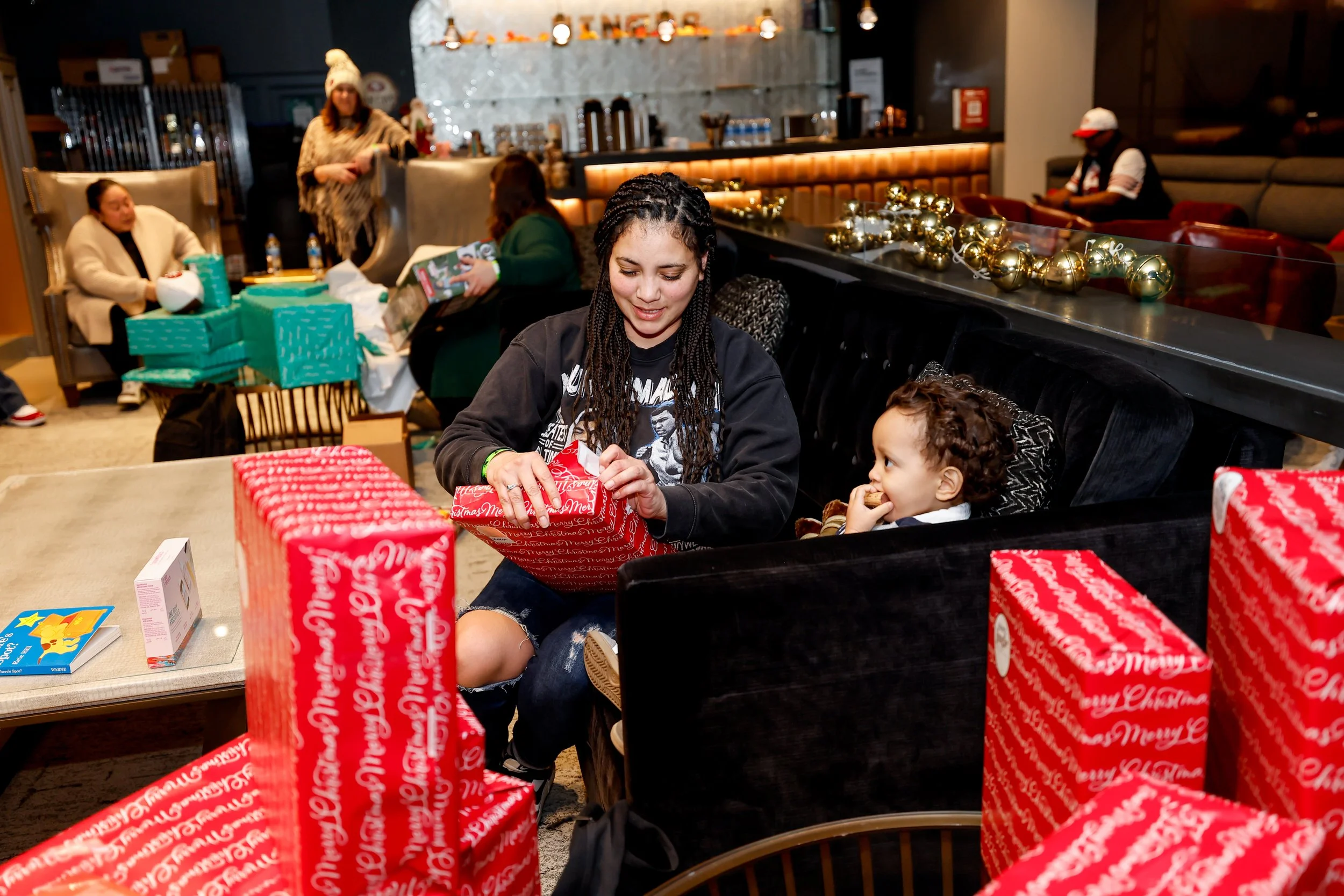 A woman with braided hair and a young girl sitting on a black couch, opening Christmas presents with red and gold wrapping. Other people are in the background at a holiday gathering, with festive decorations and gift boxes around.