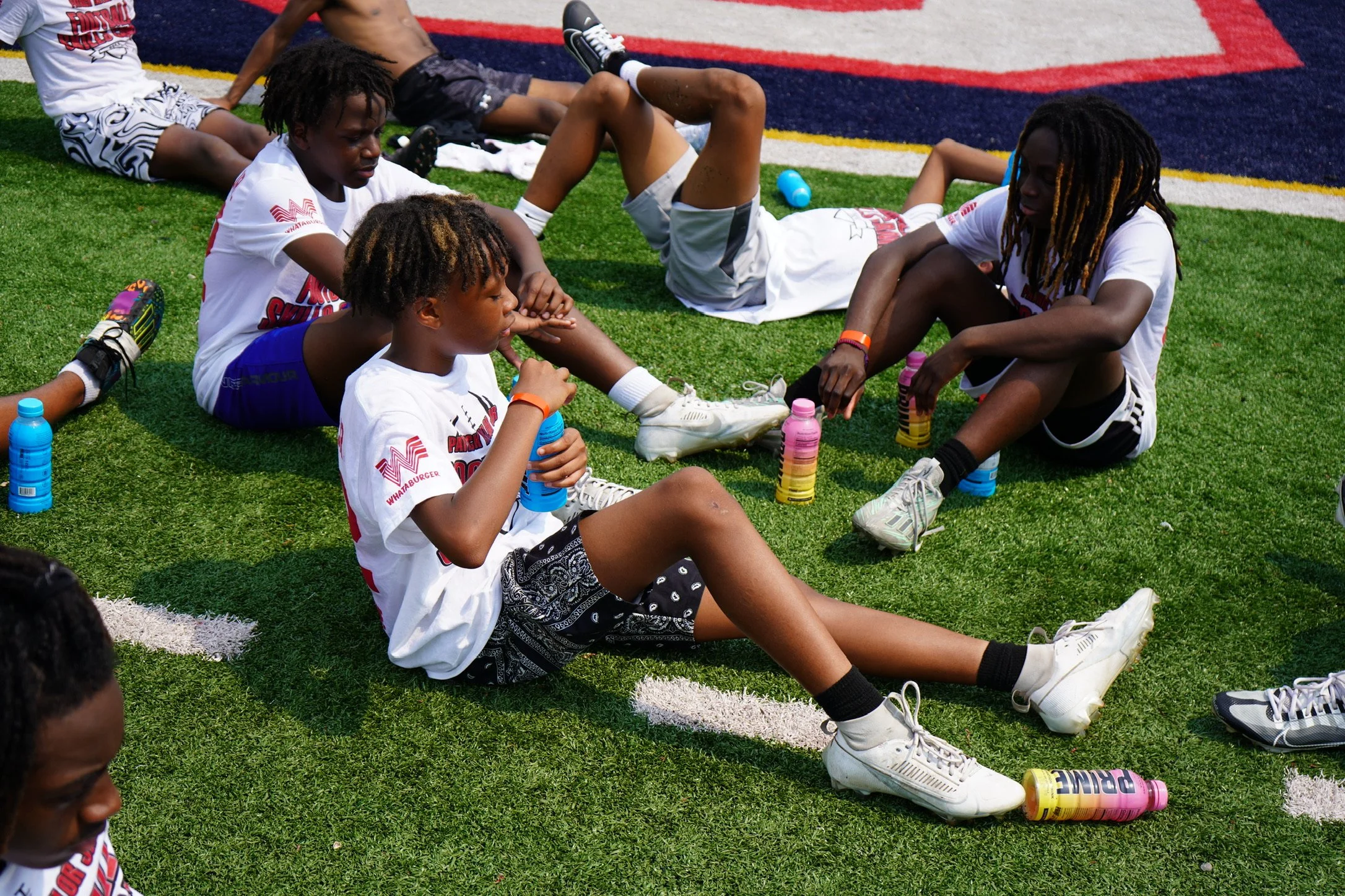 A group of young boys sitting and lying on a sports field, taking a break during practice or a game. They are wearing athletic clothing, some with water bottles around them.