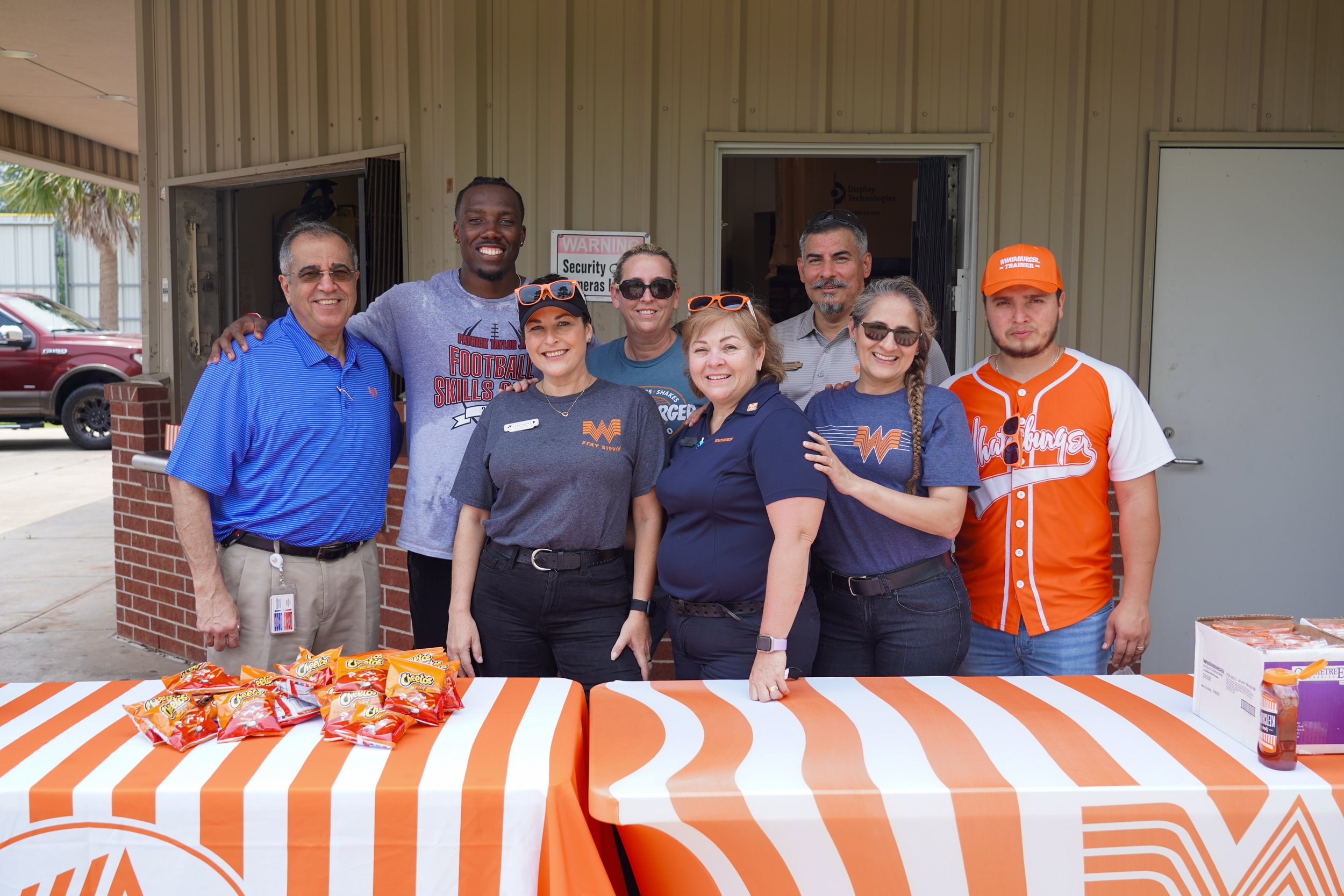 Group of eight smiling people standing behind a table with orange and white striped tablecloths, with snacks on it, outside a building with a brick wall and a gray door.