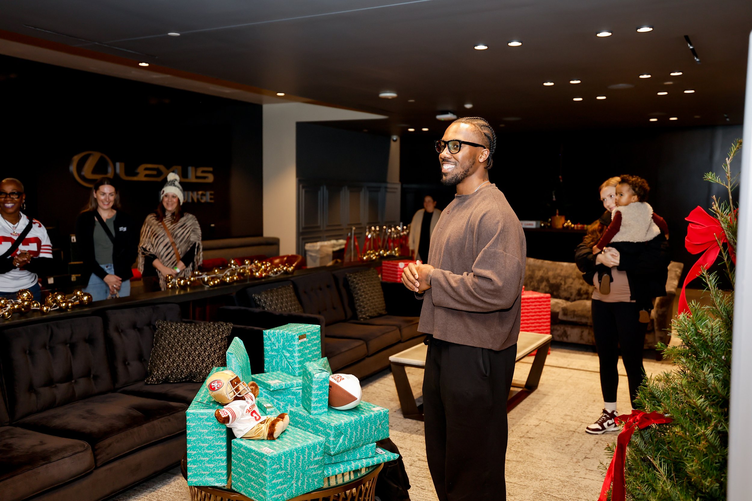 A man smiling and standing near a table with wrapped gifts and a football, with a festive Christmas setting and people in the background at the Lexus Lounge.