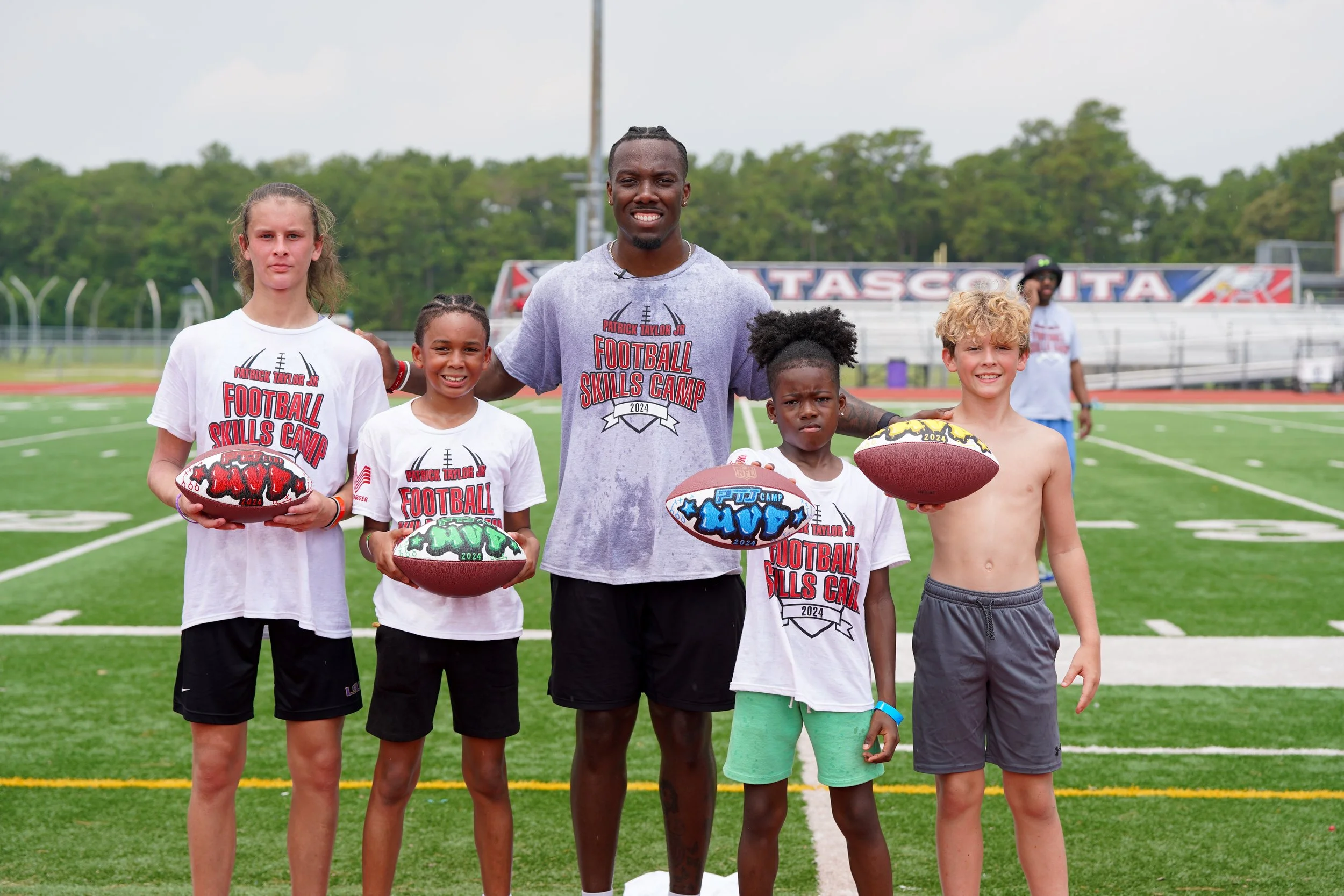 A football coach with five young players standing on a football field holding footballs, all wearing T-shirts that say 'Football Skills Camp 2024'.