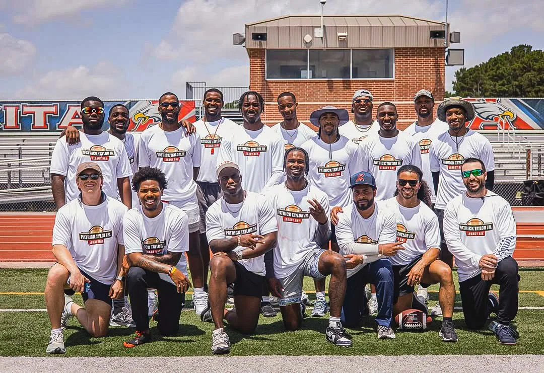 Group of young men and women in athletic gear on a sports field, posing for a team photo, some wearing sunglasses and hats, with a brick building, bleachers, and trees in the background.