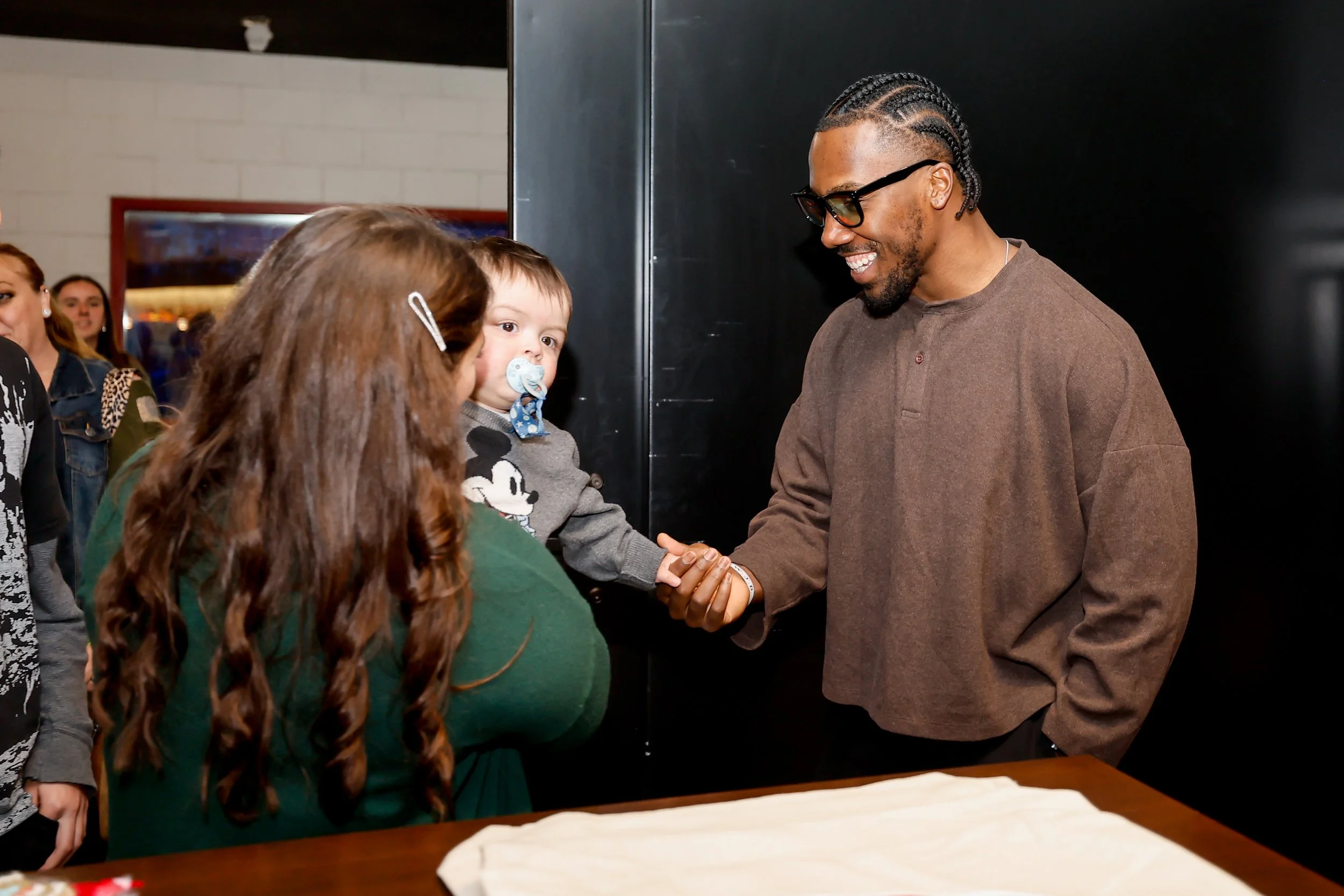 A man with glasses and braided hair shakes hands with a woman with long red hair who is holding a young boy with a pacifier, and other people are in the background.