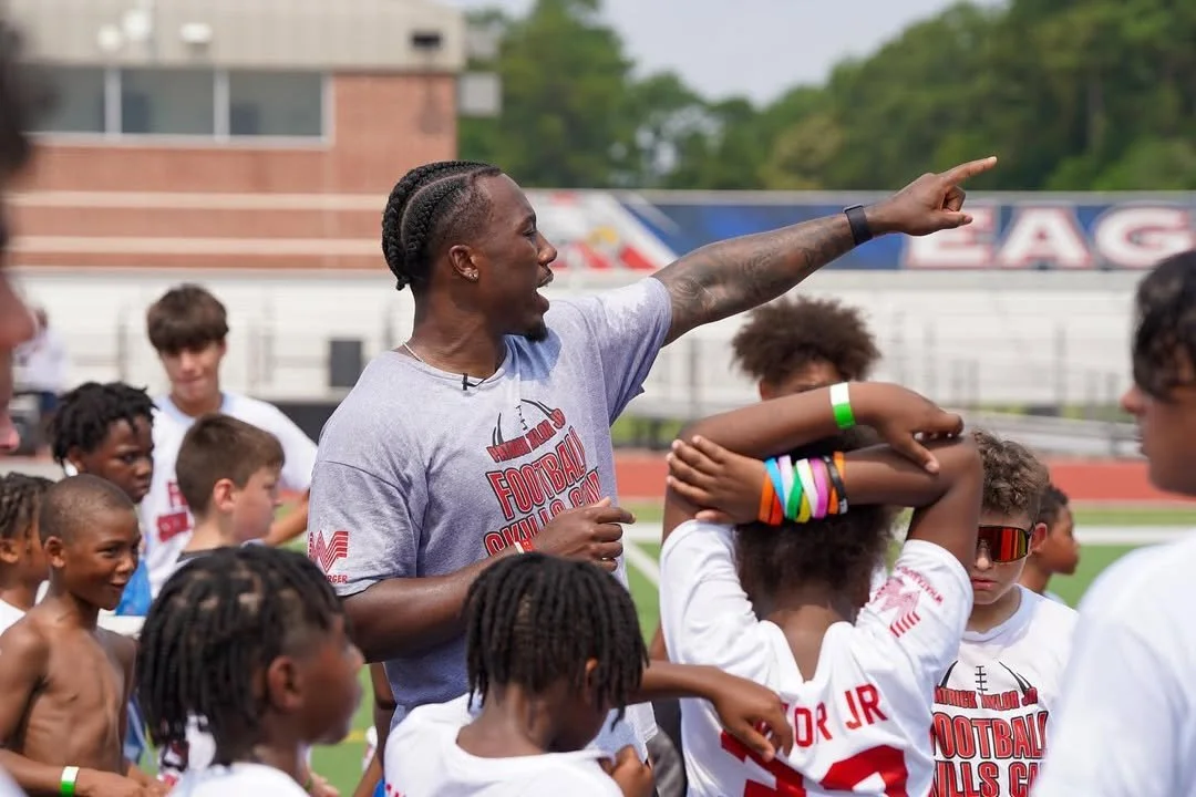 A football coach is giving instructions to young players during practice on a field, with children gathered around him, some wearing white jerseys and colorful wristbands, in an outdoor stadium.