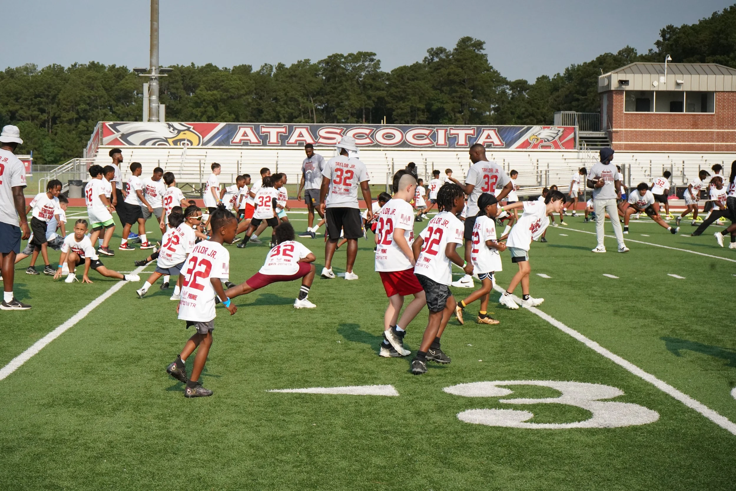 Children and coaches participating in a youth football camp on a football field, with a banner reading 'ATASSCOTA' in the background.
