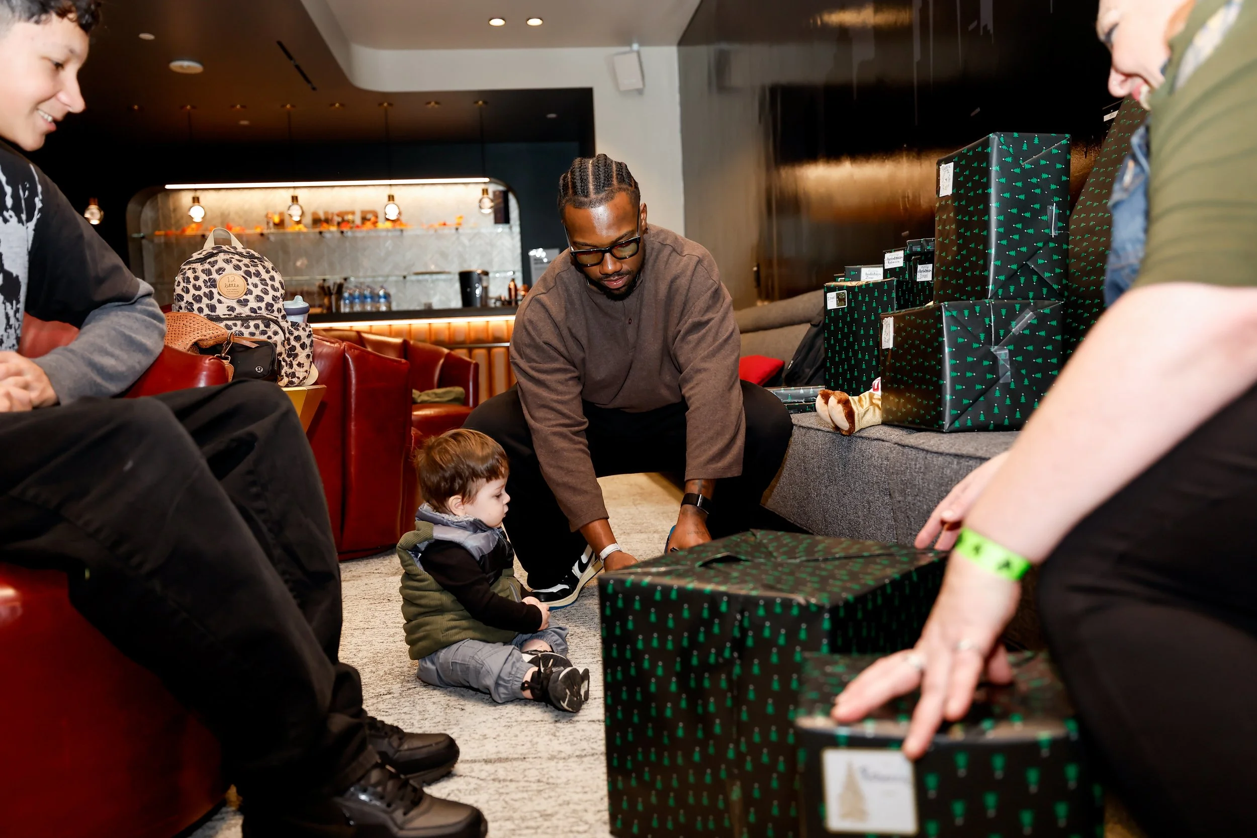 People gather around a table with wrapped presents, including a young child and adults, in a cozy indoor setting, possibly for a holiday gift exchange.