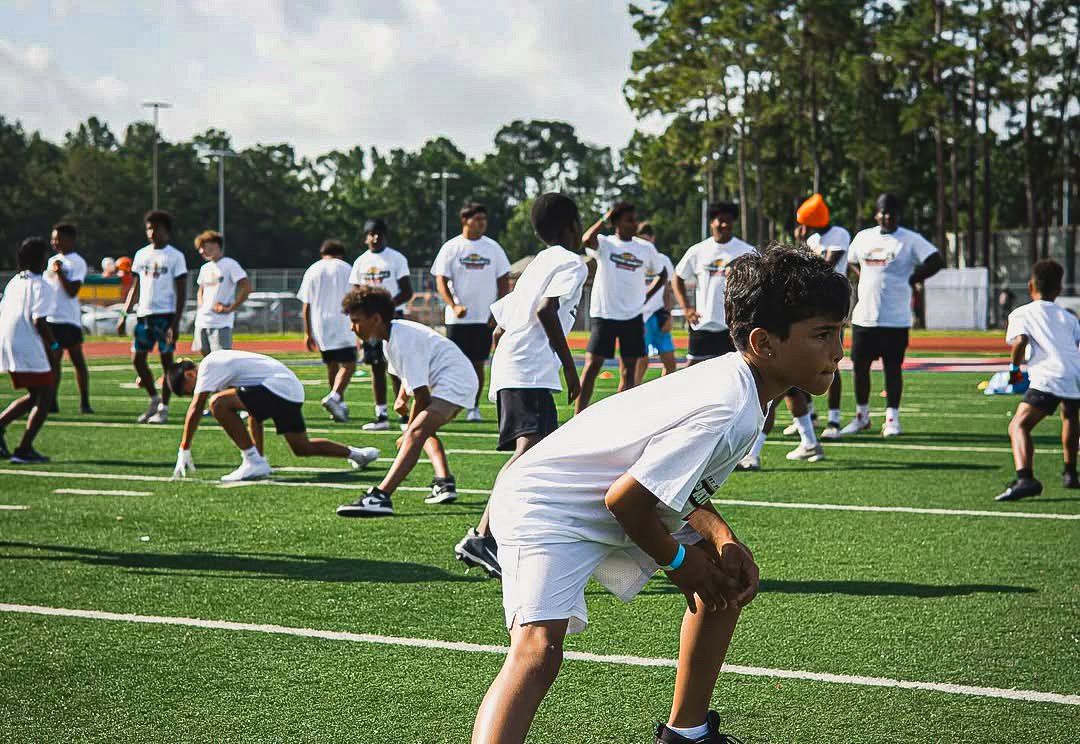 Children participating in a sports practice on a green field, some in a crouched position, many wearing white T-shirts and shorts.