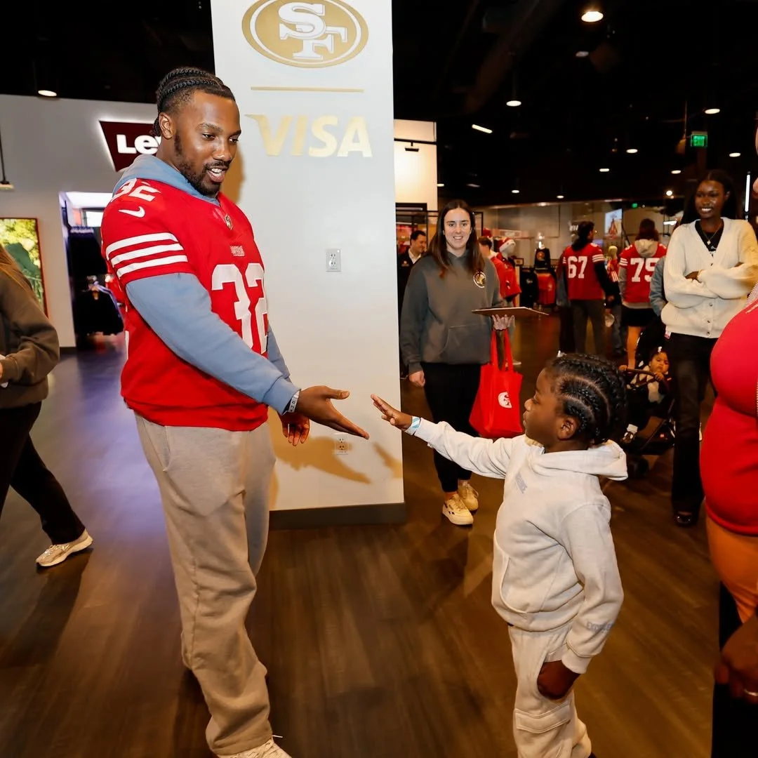 giving a fist bump to a young girl in a gray sweatshirt and matching pants inside a sports store. Other people are in the background, including women wearing 49ers jerseys, one holding a tablet, and logos for Levi's and Visa are visible on the wall.