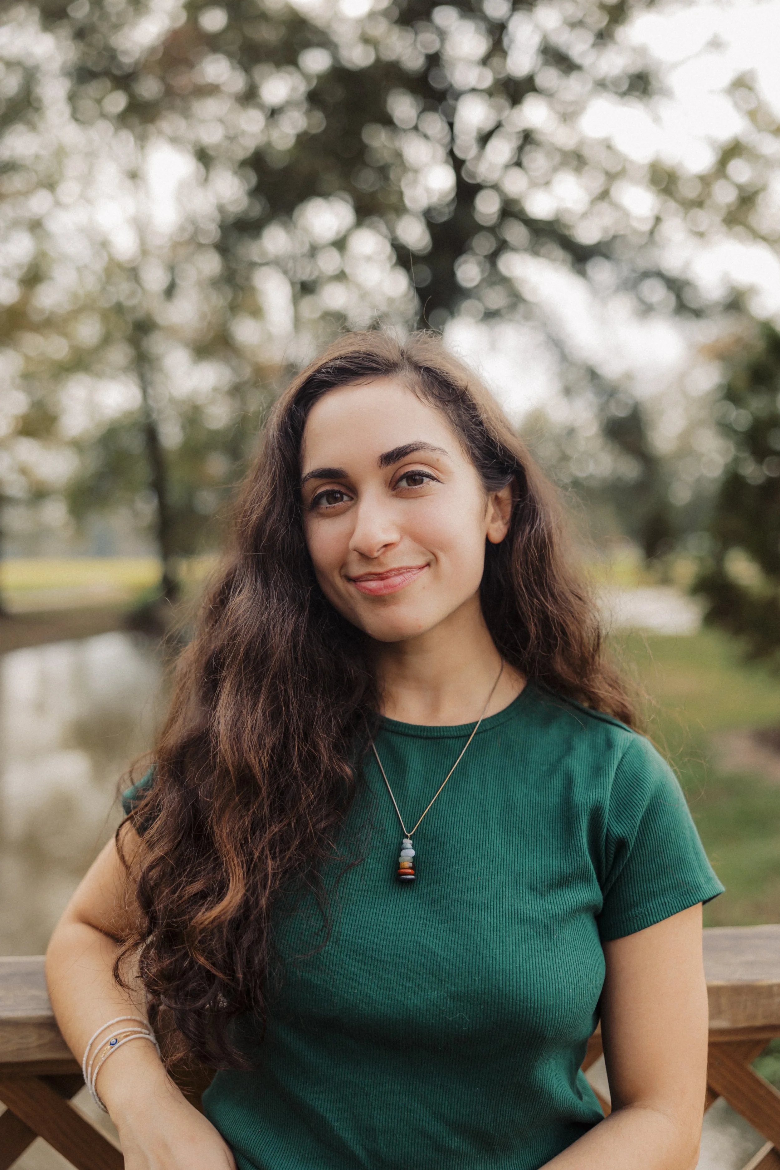A woman with long curly brown hair wearing a green shirt and a beaded necklace, standing outdoors near a wooden railing with trees and a pond in the background.