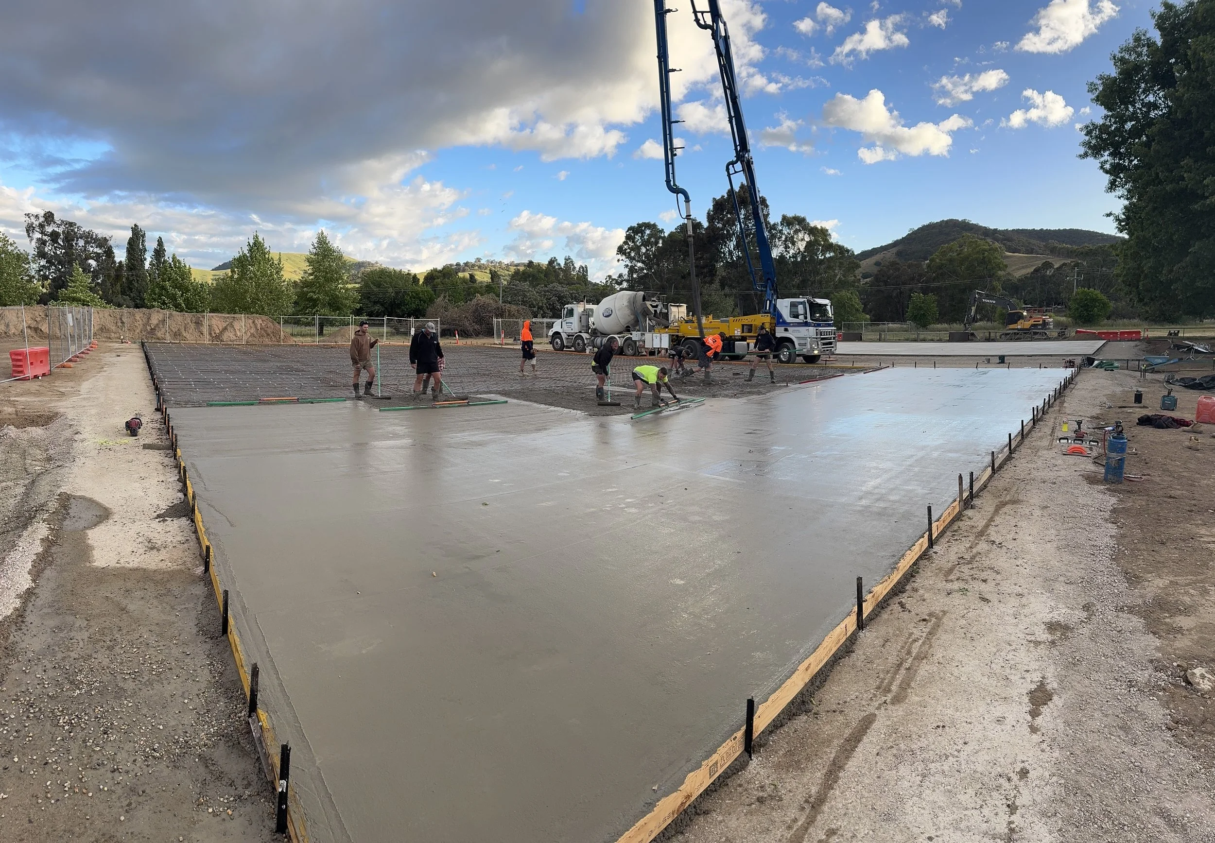 Workers pouring and leveling concrete on a construction site for a building foundation with hills and trees in the background under a partly cloudy sky.