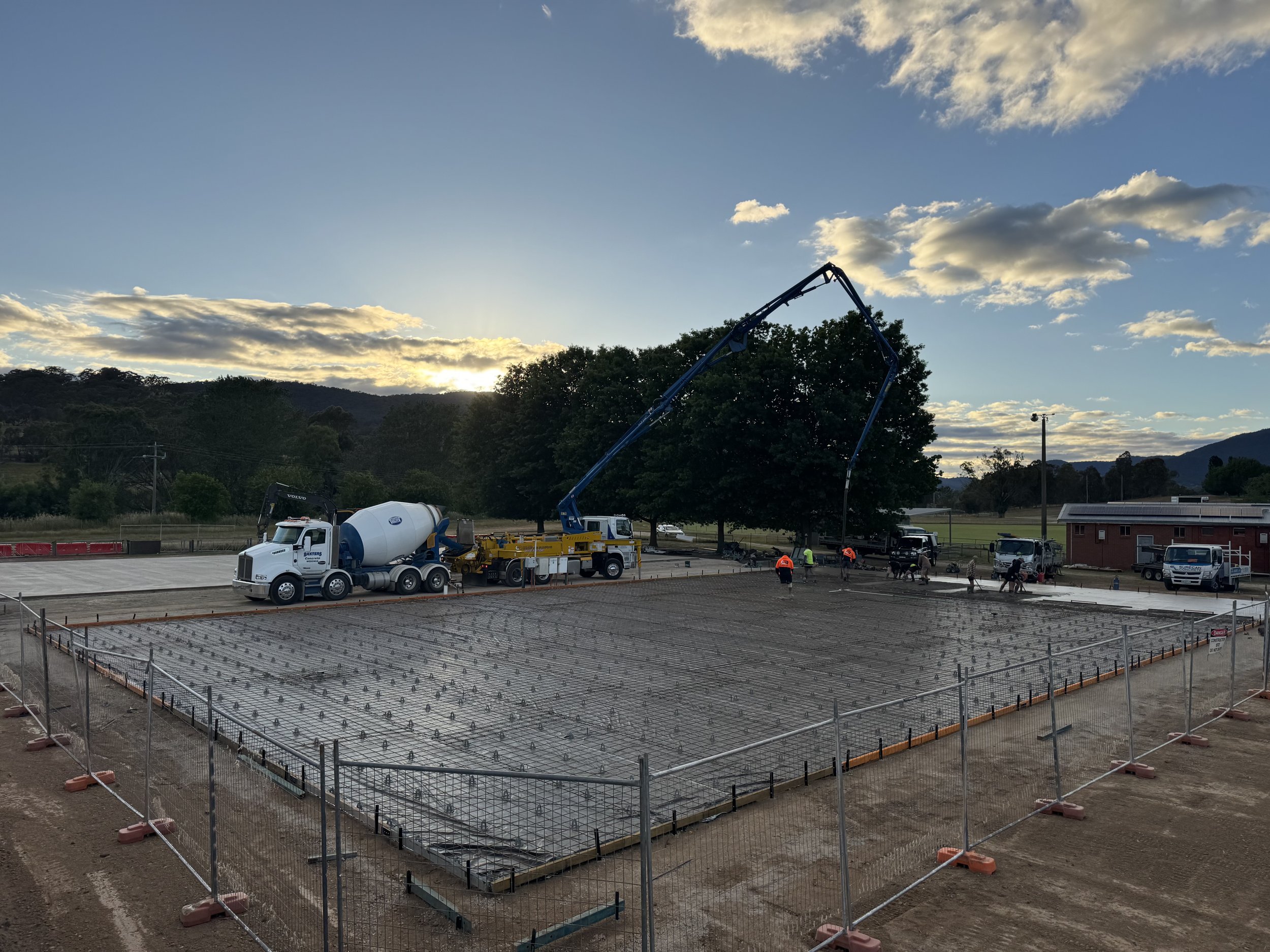 Construction site with workers pouring concrete on a foundation, using a concrete mixer truck and concrete pump, with a tree and mountains in the background at sunset.
