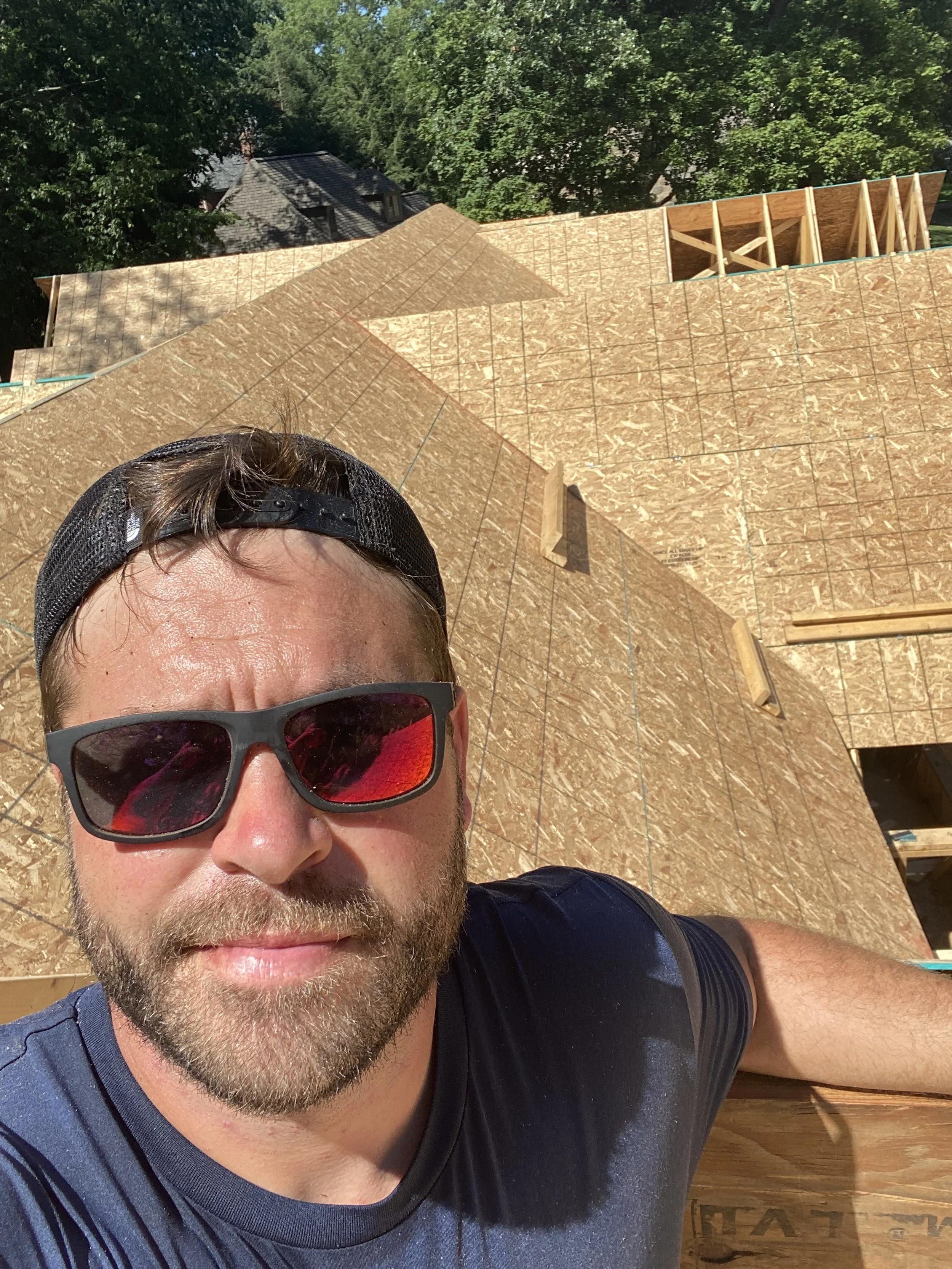 A man wearing sunglasses and a backward cap takes a selfie at a construction site with a house being built, showing wooden framing and roof sheathing.