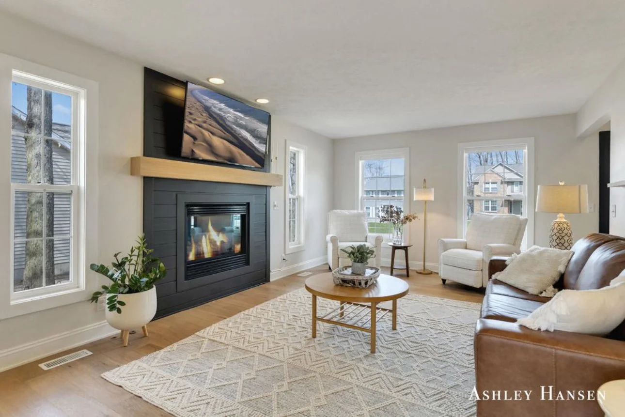 Bright living room with a fireplace, flat-screen TV above, two white armchairs near windows, a brown leather sofa, a coffee table with a plant, and several lamps, with a patterned rug on hardwood floor.