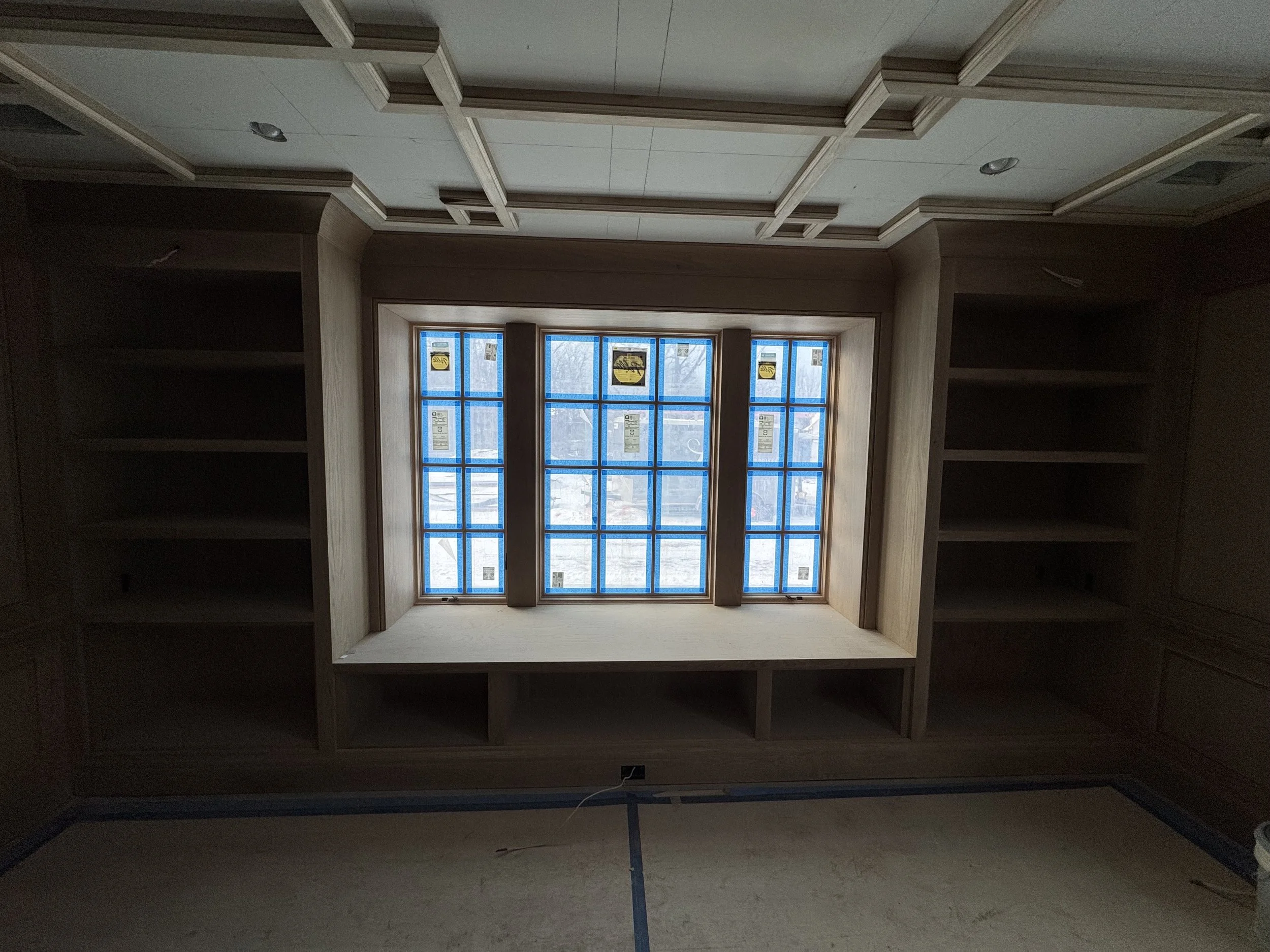 Unfinished built-in wooden shelves around a large bay window with blue painter's tape in a room under construction.