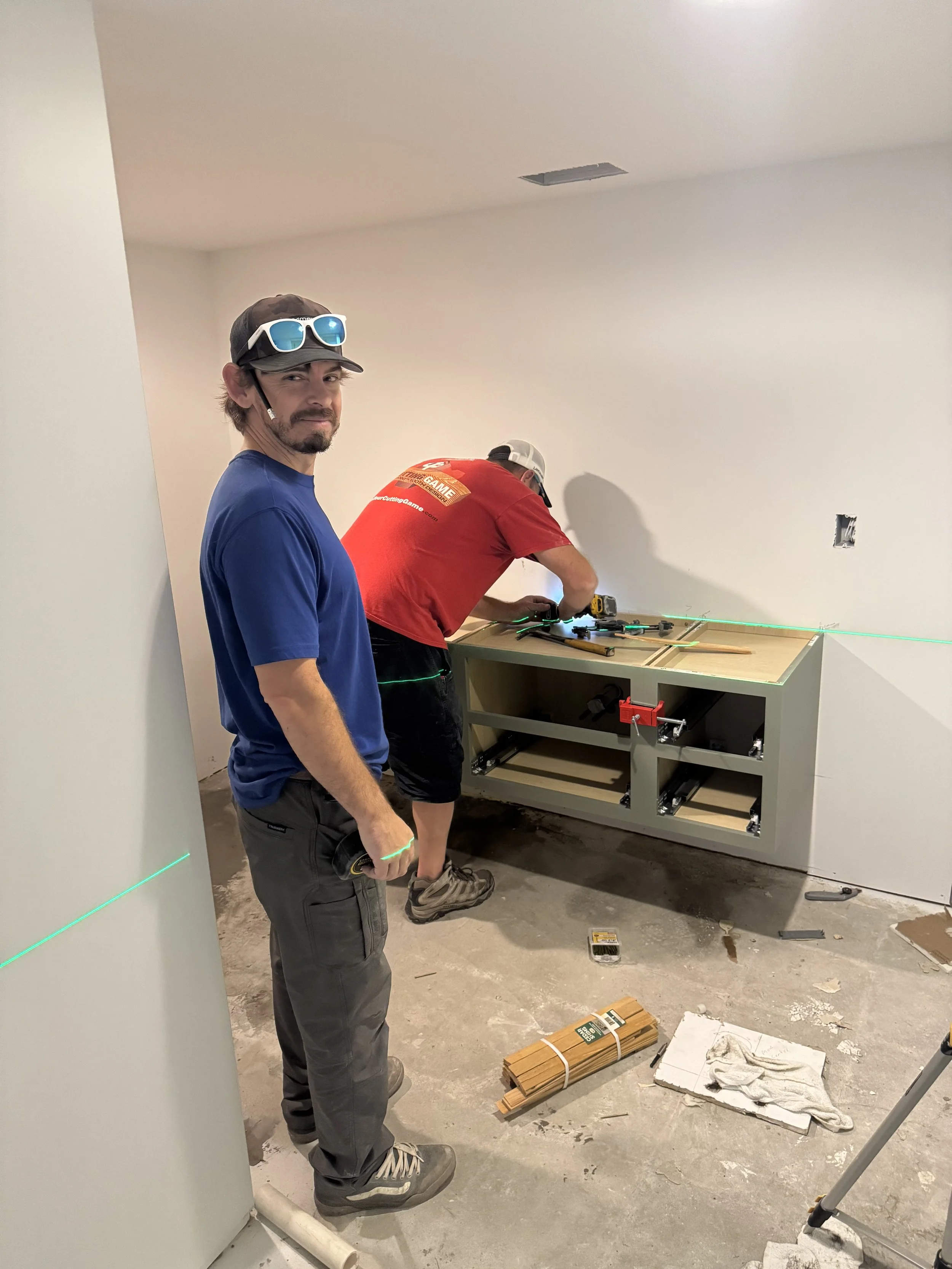 Two men working on a construction project in a room with white walls. One man is facing the camera, wearing a blue shirt, gray pants, sunglasses on his head, and holding a measuring tape. The other man, wearing a red shirt and a cap, is bent over a partially assembled piece of furniture, using tools. There are laser levels, tools, and construction materials on the floor.