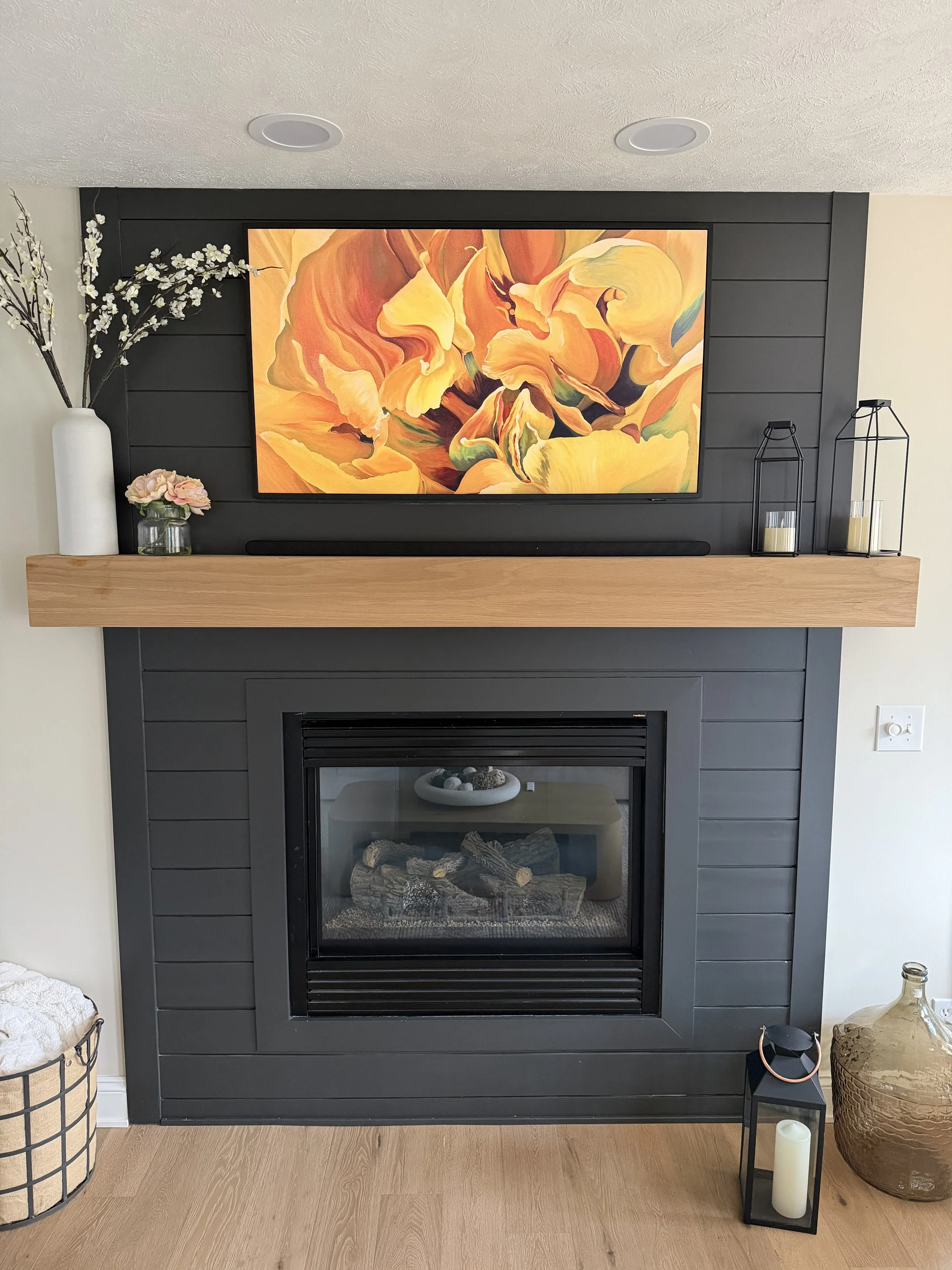 Living room fireplace with a black wooden panel surround, a wooden mantel, and a colorful abstract floral artwork above. Decor includes vases with flowers, lanterns with candles, and a large vase on the floor.