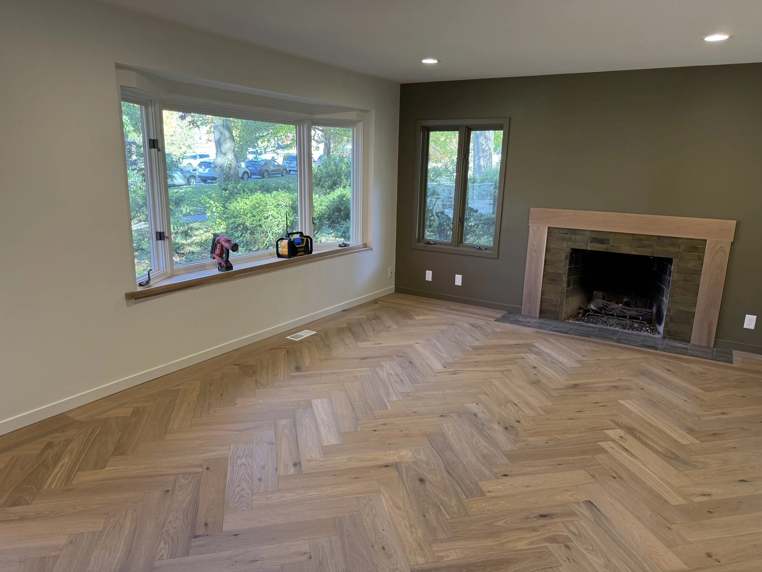 Empty living room with hardwood floor, gray accent wall with fireplace, two windows, tools on window sill, and ceiling lights