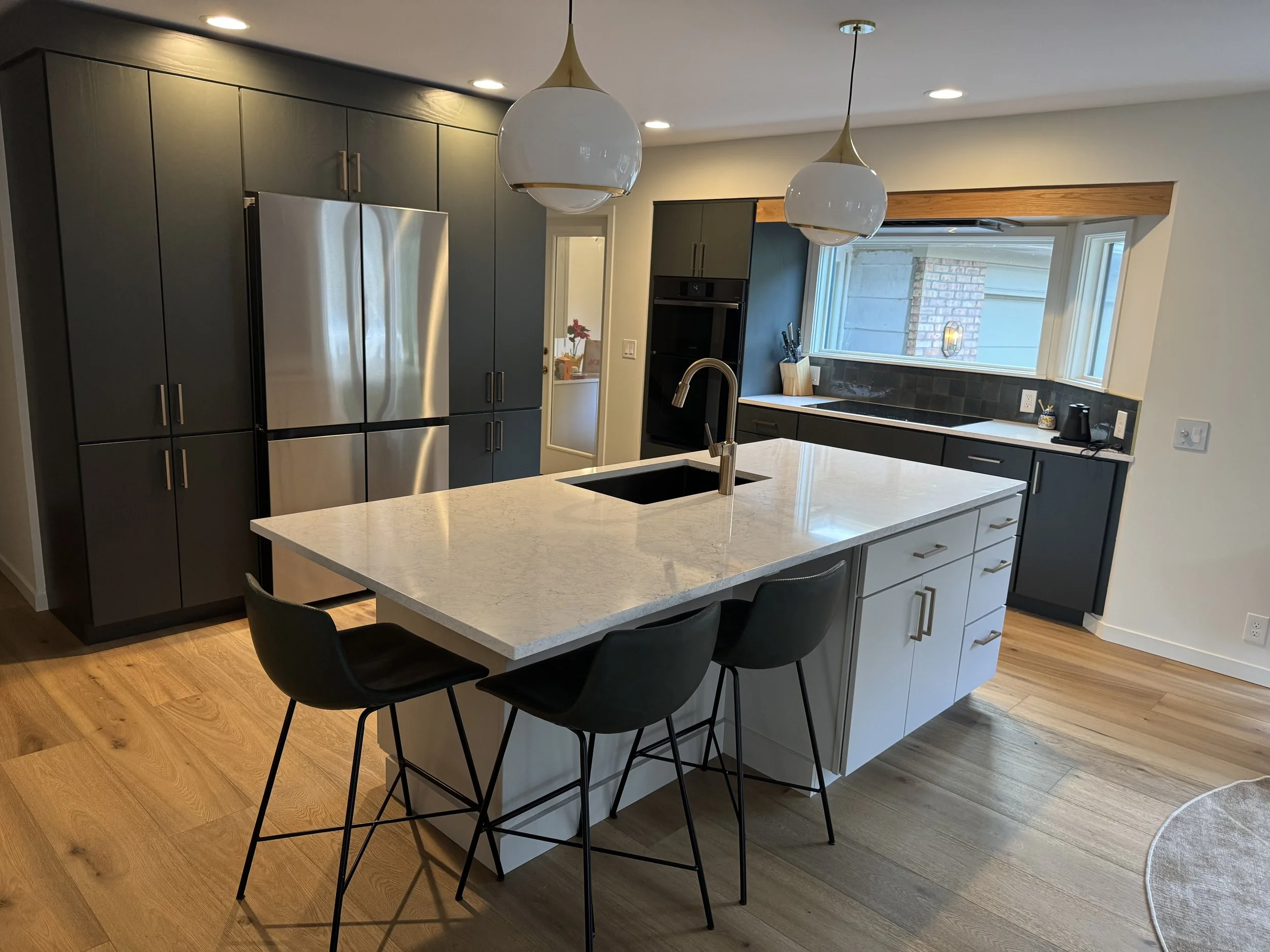 Modern kitchen with a large white island countertop, black bar stools, stainless steel refrigerator, dark cabinetry, and a window overlooking the outdoors.