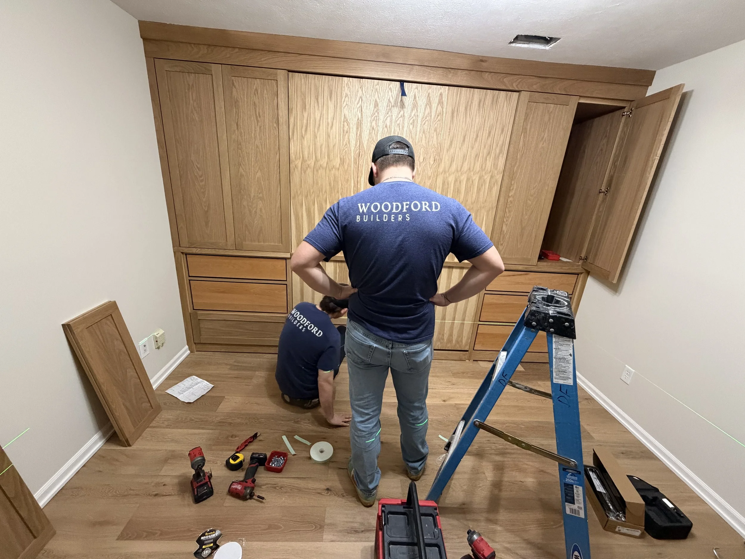 Two workers from Woodford Builders installing a Murphy bed cabinet in a basement bedroom, one standing and one kneeling, with tools and hardware on the floor.