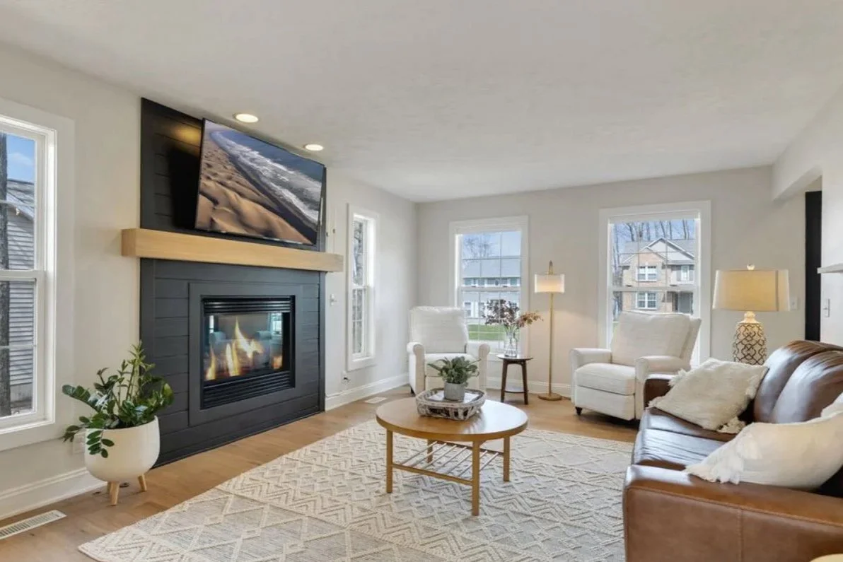 Living room with sofa, armchairs, fireplace, mounted TV, side tables, lamps, and large windows, decorated in neutral tones.