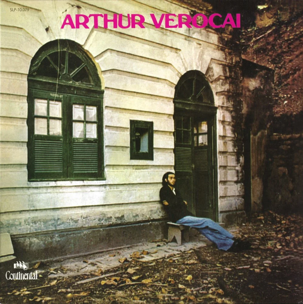 Album cover titled "Arthur Verocai" featuring a man sitting on a bench against a building with two arched windows and a door, surrounded by fallen leaves and trees. The top of the cover has pink text with the artist's name, and the bottom left corner