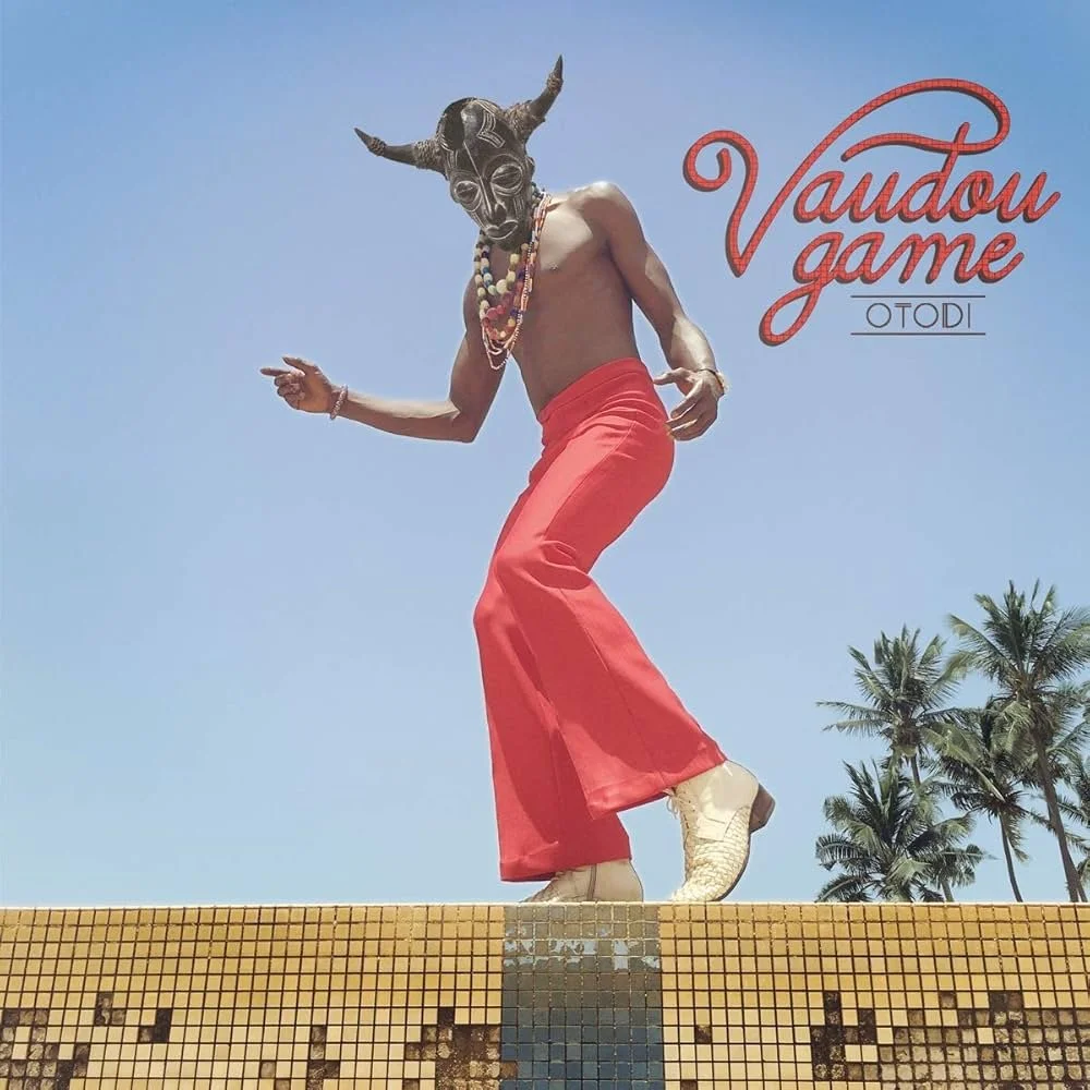 A person wearing a traditional mask, red pants, and white shoes is dancing outdoors on a tiled surface with palm trees in the background, under a clear blue sky, with the words 'Vaudou game' and 'OTODI' written in red on the image.