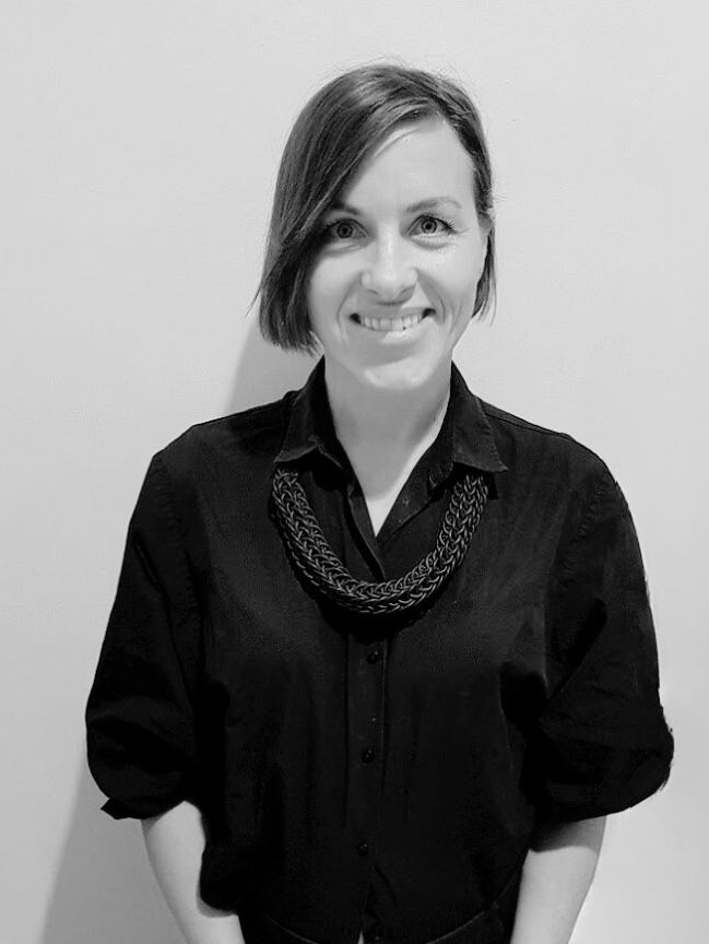 Black and white photo of a smiling woman with short hair, wearing a dark shirt and a chunky scarf, standing against a plain wall.