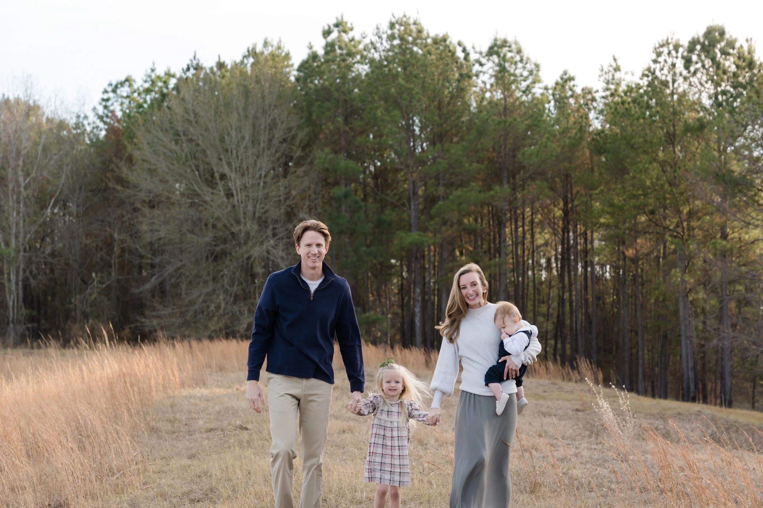 A family of four walking outdoors in a grassy field with trees in the background during fall, smiling and holding hands.