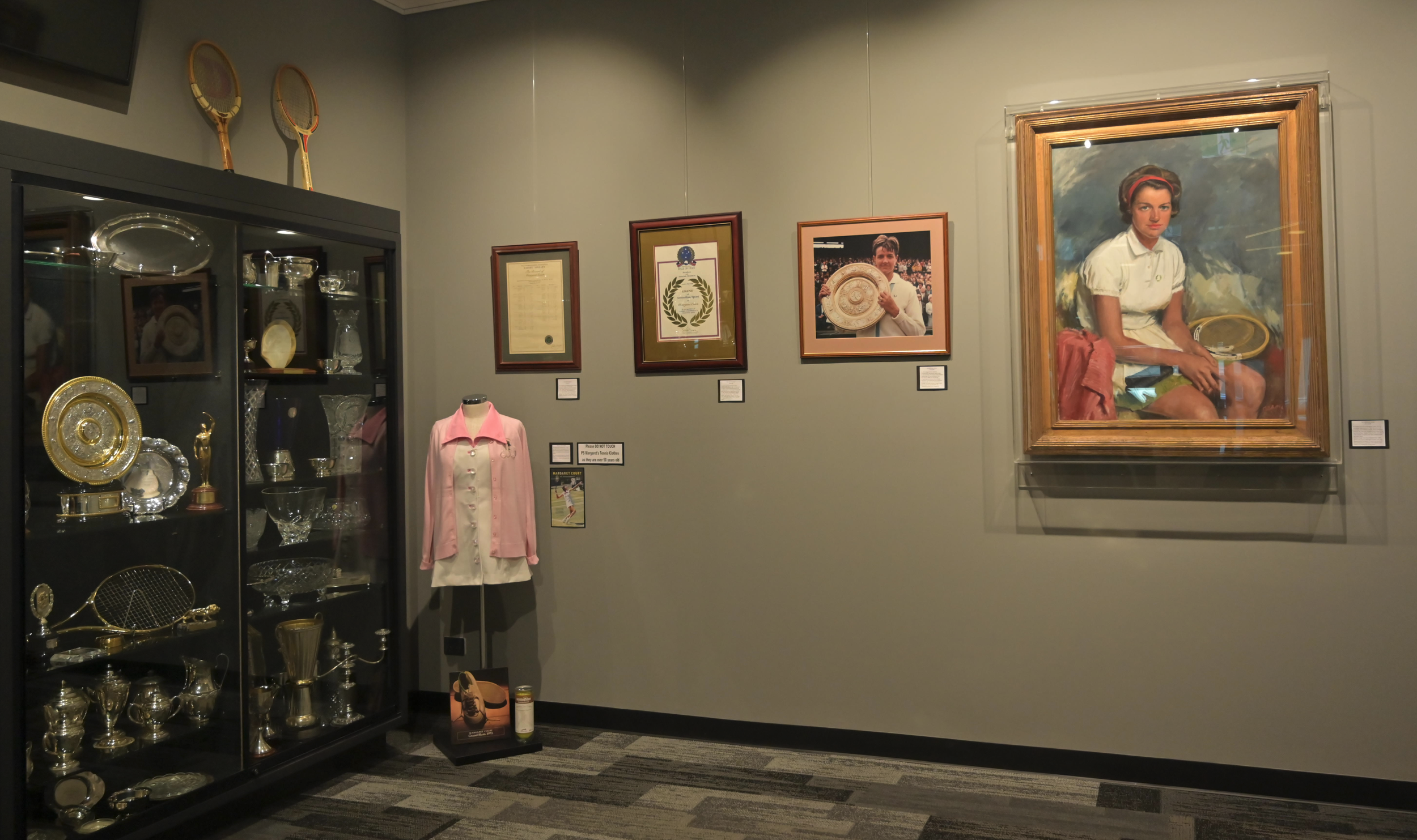 Exhibit of tennis trophies, a framed photograph of a woman holding a trophy, a large portrait of a woman with a tennis racket, and a glass case with glassware and awards in a museum.