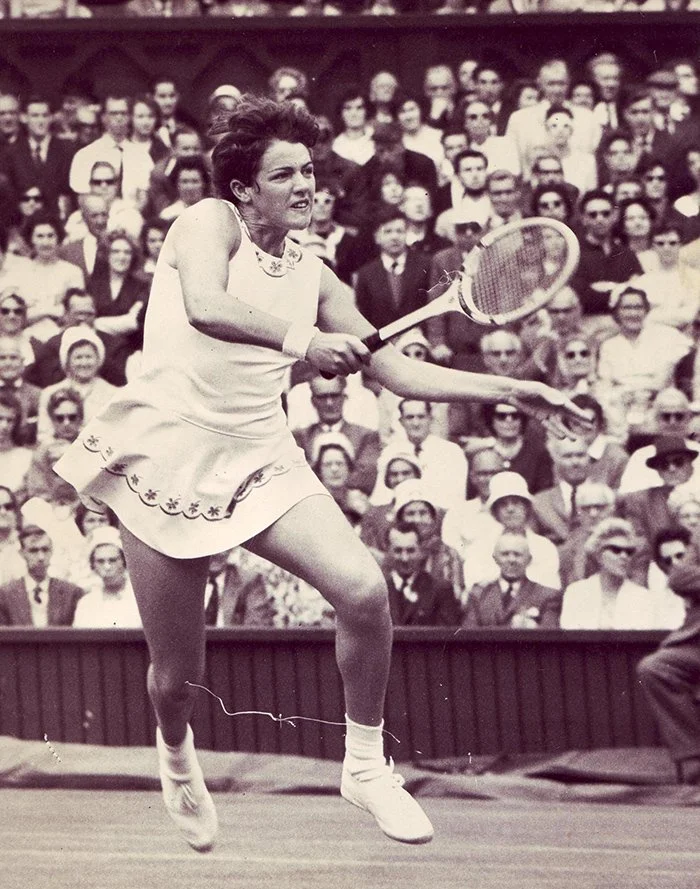 Black and white photo of a female tennis player in a sleeveless dress mid-swing during a match, with a large crowd of spectators seated in the background.