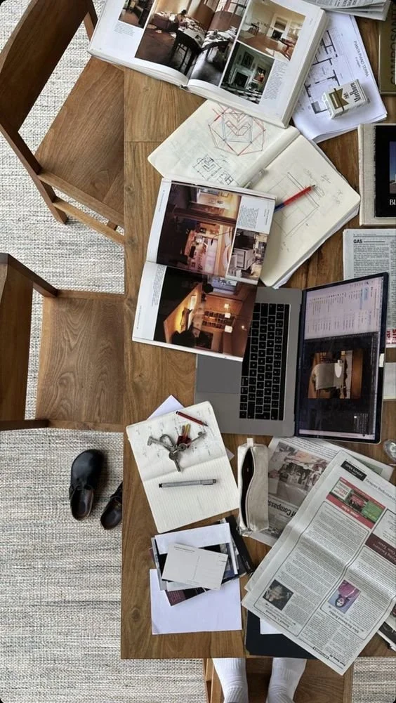 Overhead view of a wooden dining table cluttered with open magazines featuring interior design and architecture, a laptop displaying a website, notebooks with sketches and notes, a pen, keys, a pair of black shoes, and a newspaper. A person wearing white socks is standing in front of the table.