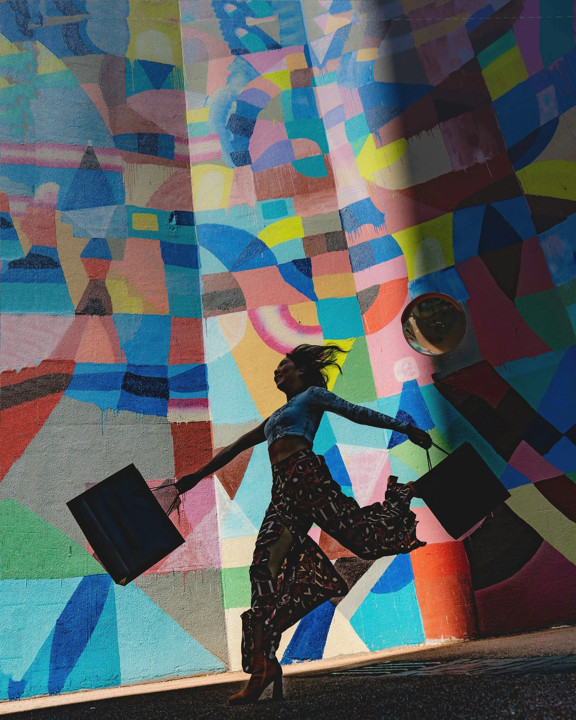 A woman with long hair, wearing high heels, a blue patterned top, and wide-legged patterned pants, is jumping in front of a colorful abstract mural while holding shopping bags in both hands.