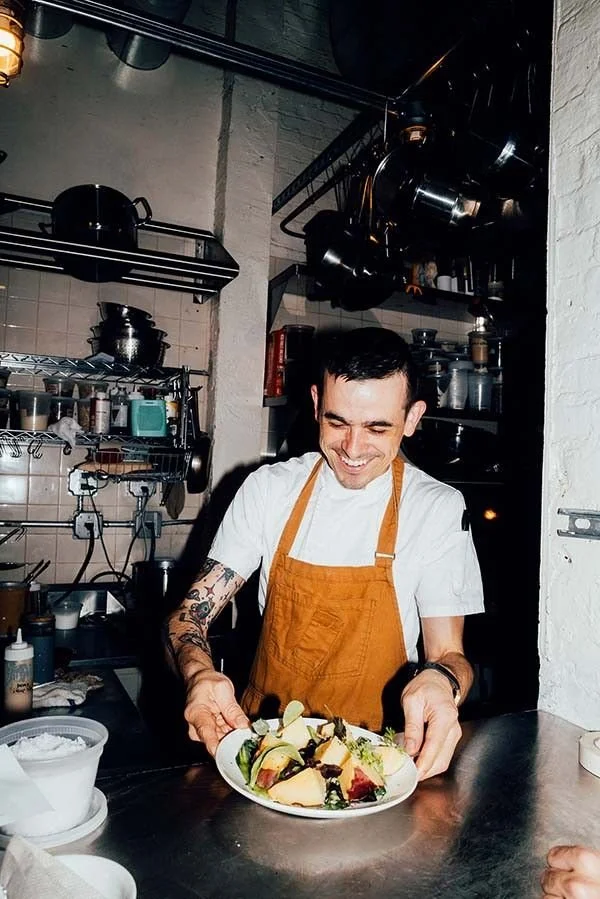 A smiling chef with tattoos, wearing a white shirt and orange apron, presenting a colorful salad in a restaurant kitchen.